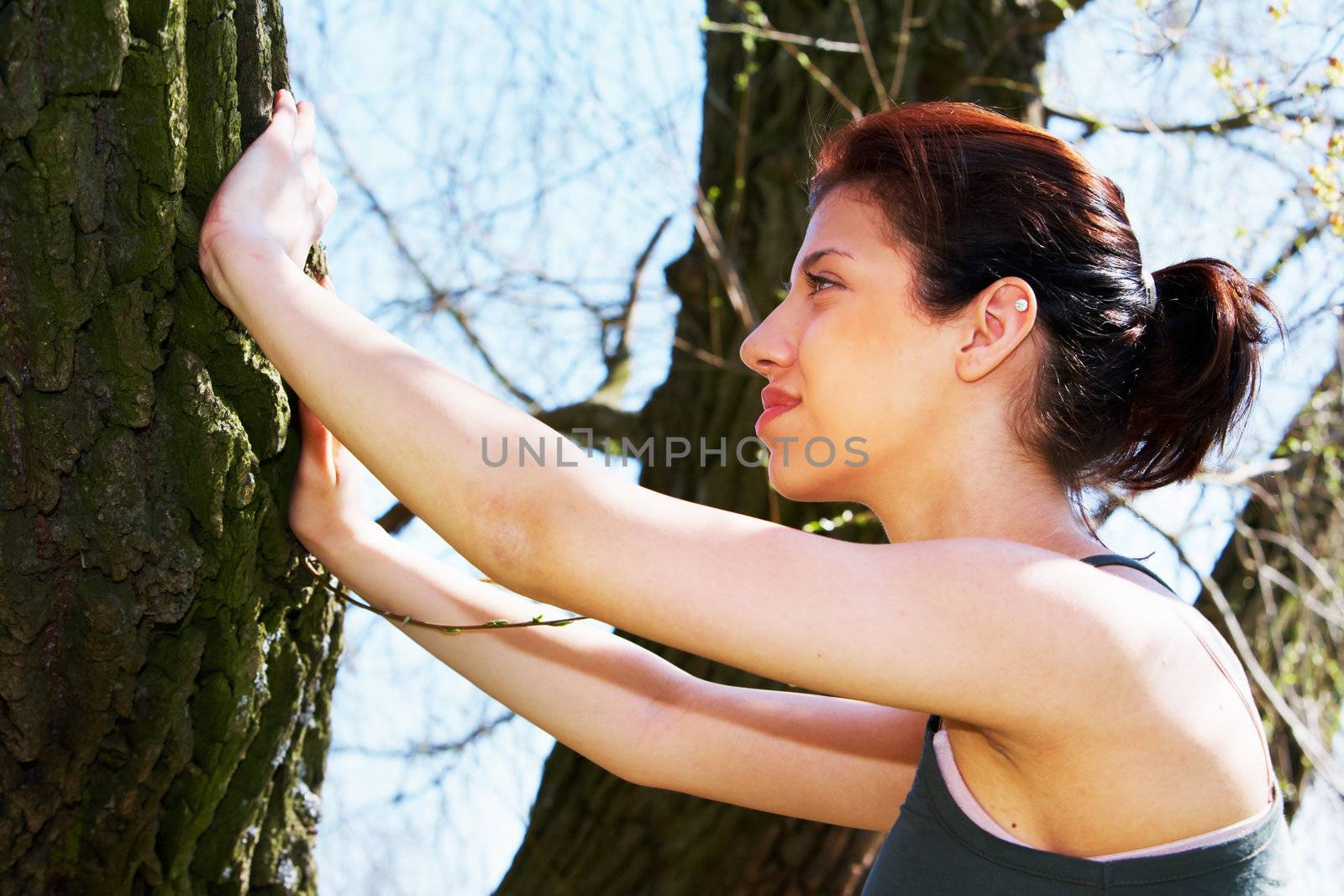 Jogging teenage girl stretching using tree, side profile