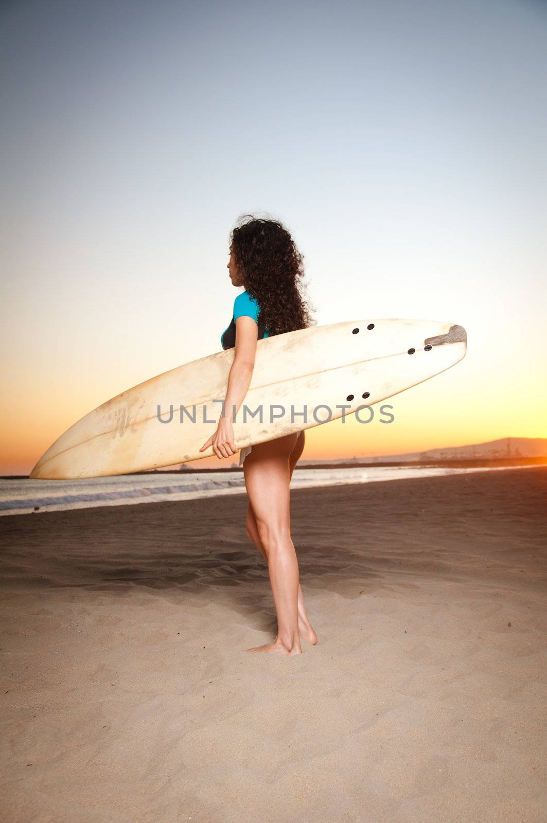 Beautiful model wearing a white bikini and surfer outft holding a surfboard during sunset by the beach