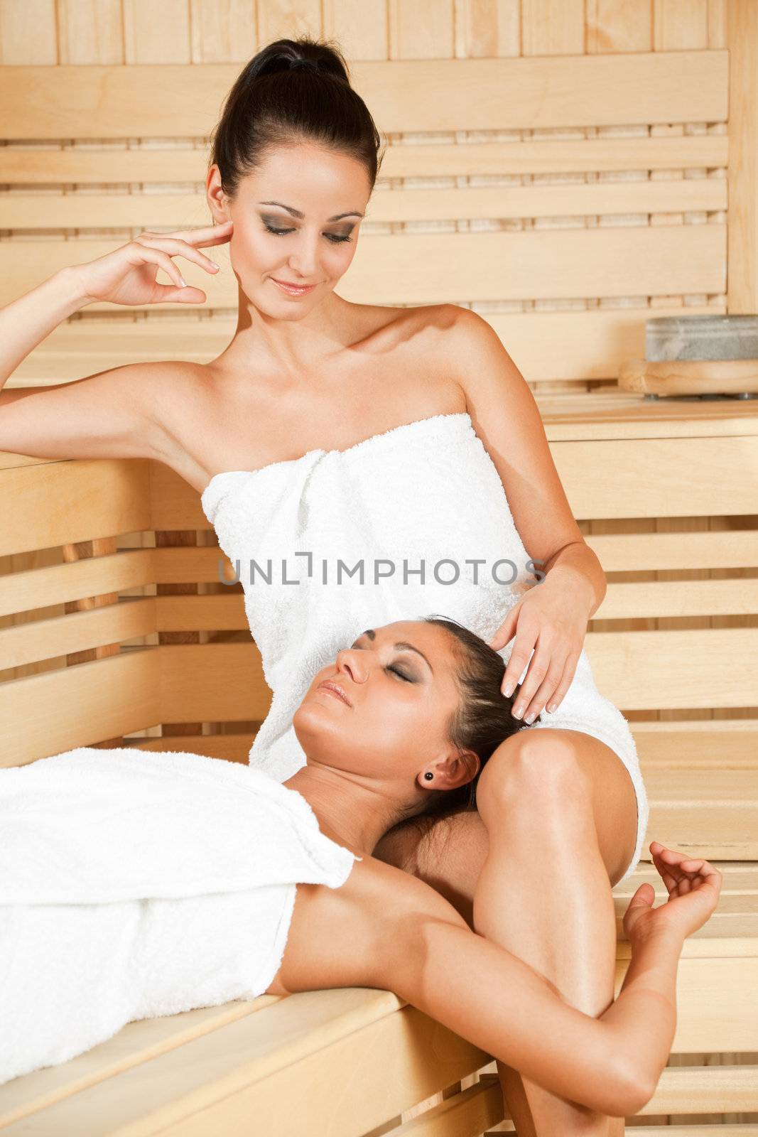 Two beautiful females relaxing at sauna, one lying in other's lap