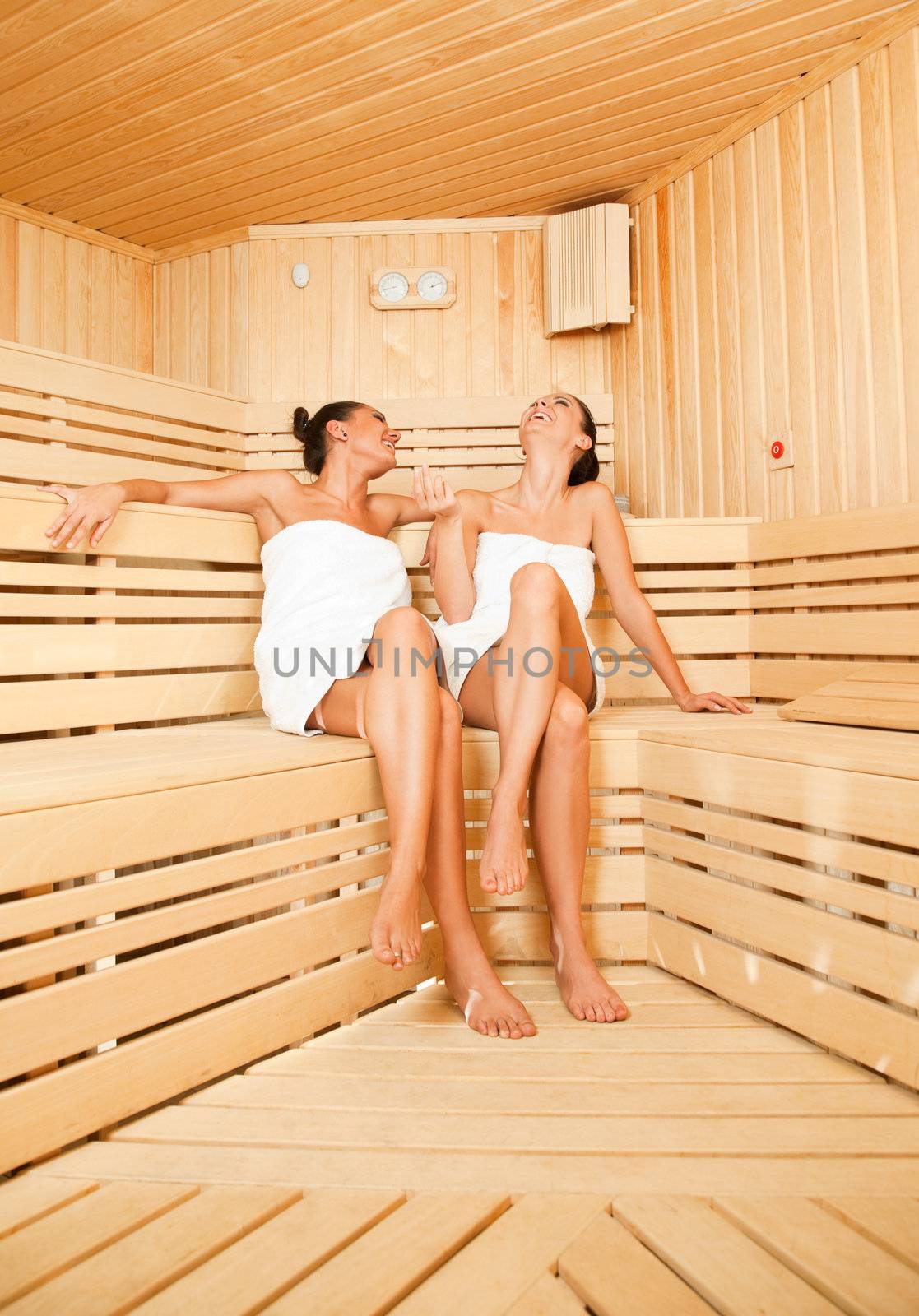 Two beautiful females with towels relaxing at sauna chatting and laughing