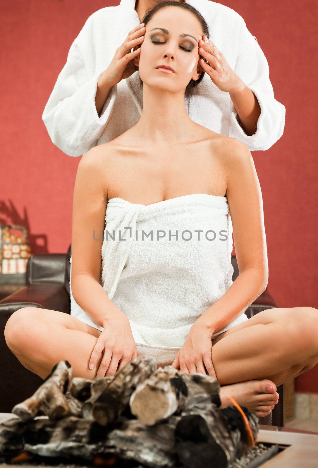 Young female sitting with legs crossed behind fireplace, receiving facial massage