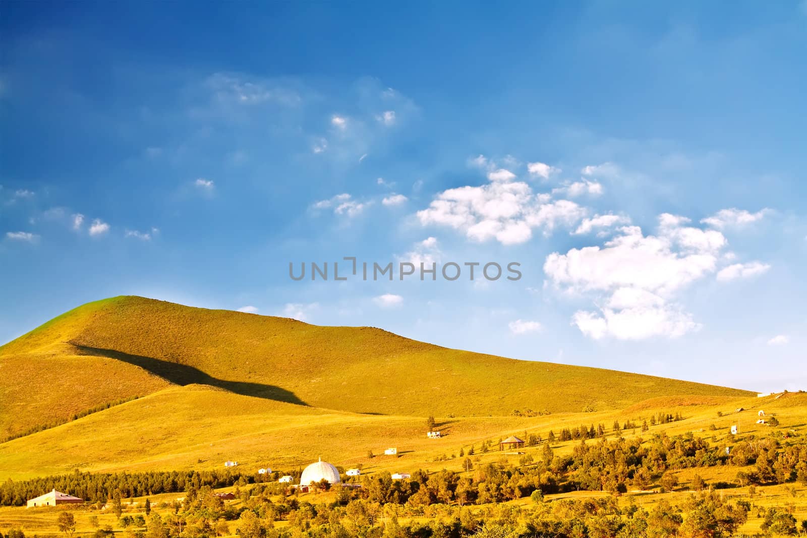 Landscape photo of plain with colorful grass under blue sky, with small huts aside a road along the hill