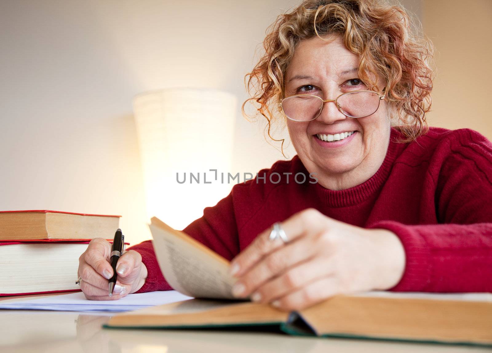 mature female is sitting by a table with books and a pen, smiling