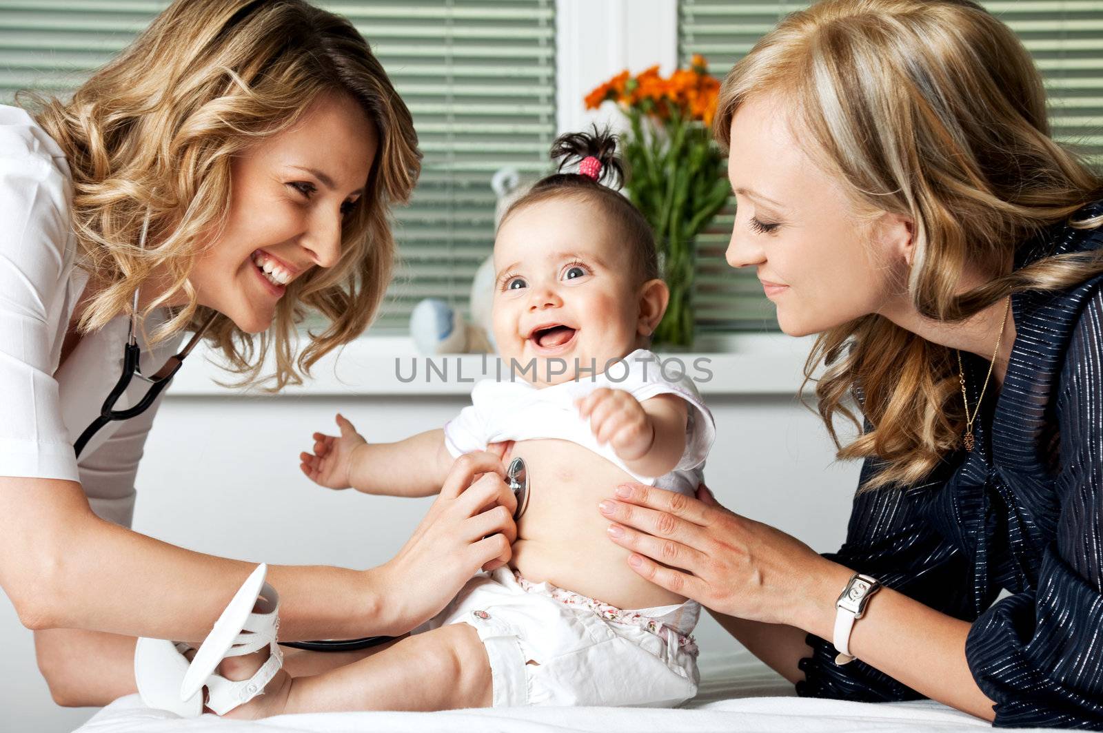 Beautiful female doctor examining little smiling baby girl, held by mother