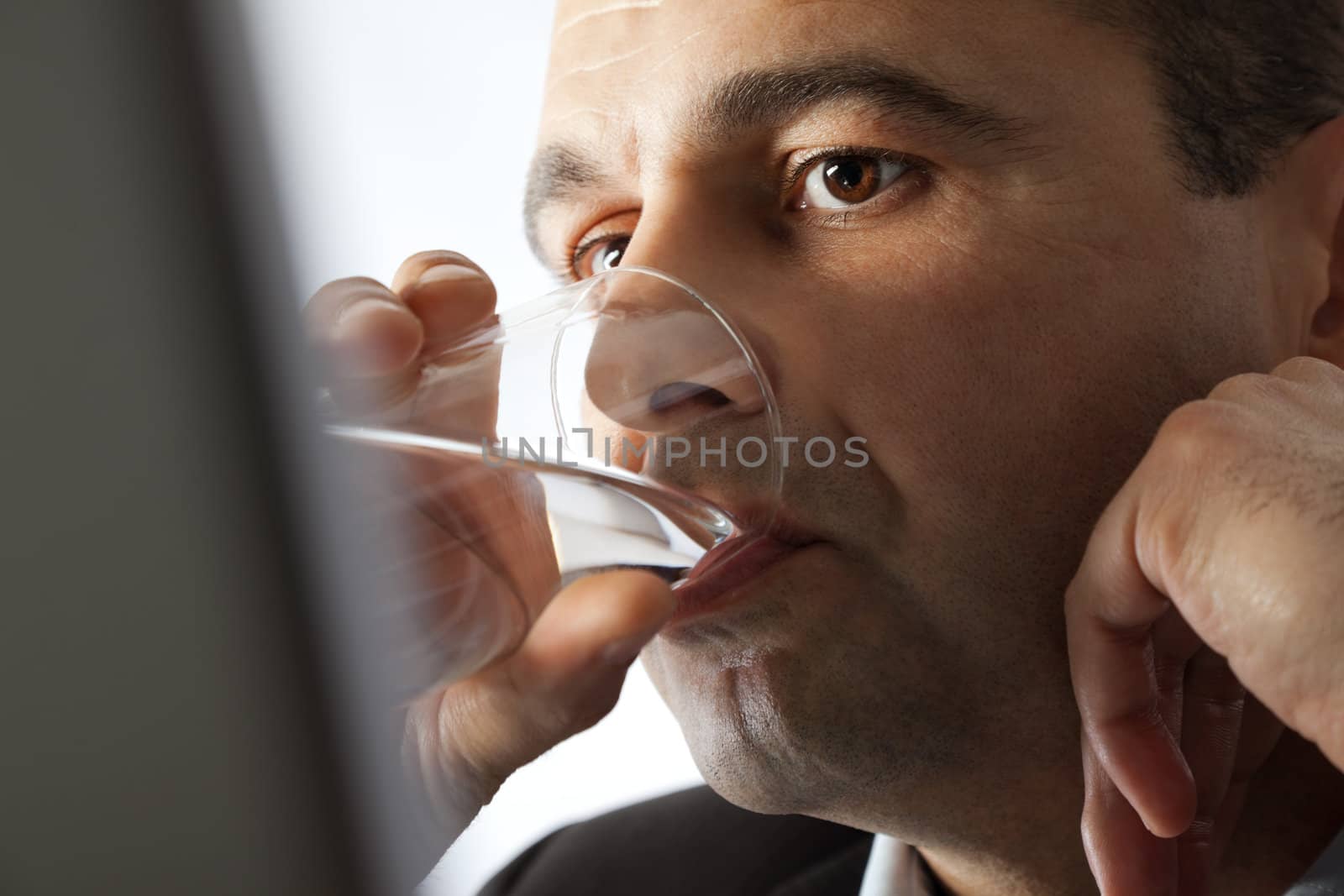 Close-up of man's face drinking water from a glass, behind laptop