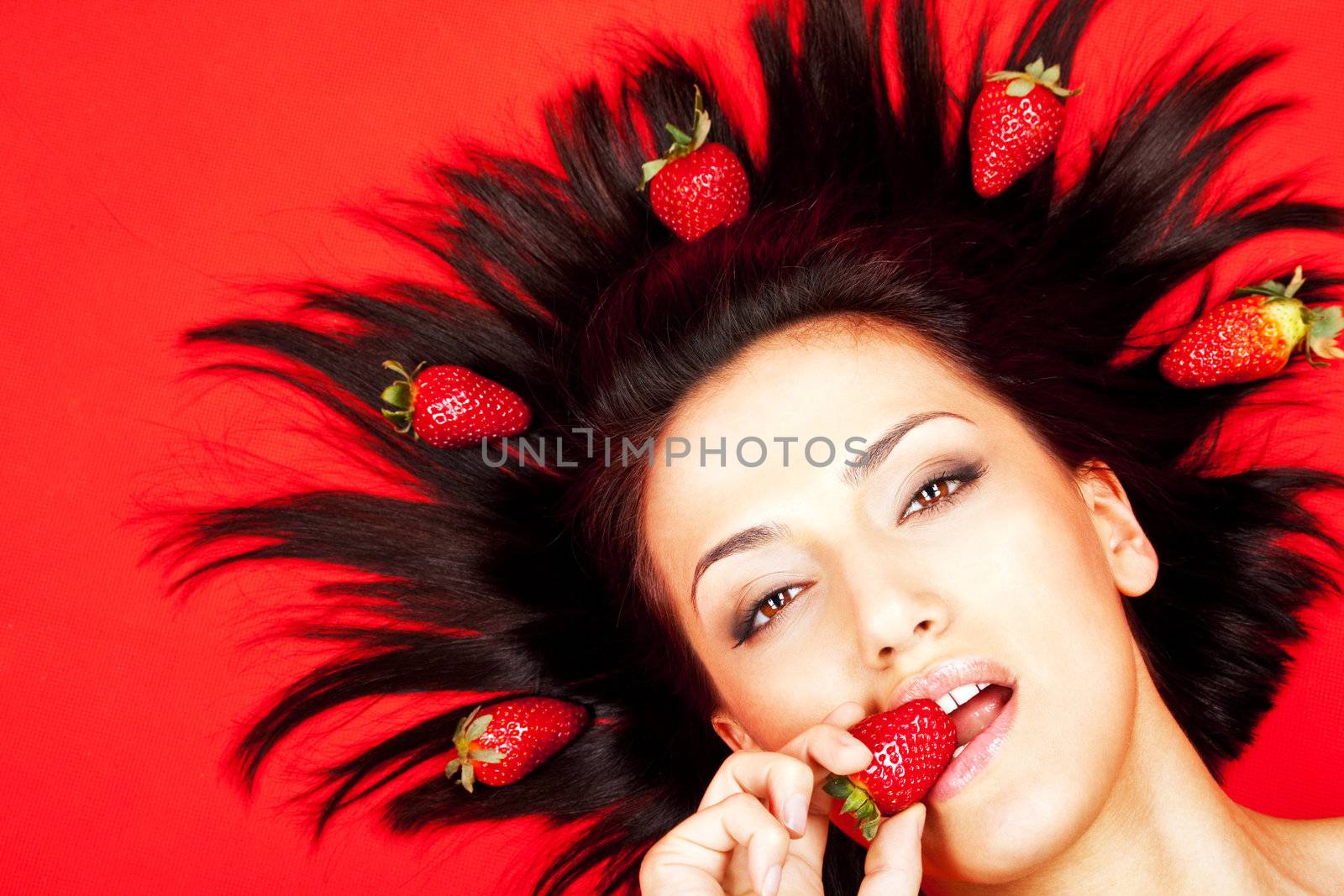 Portrait of woman lying, biting strawberry on red background