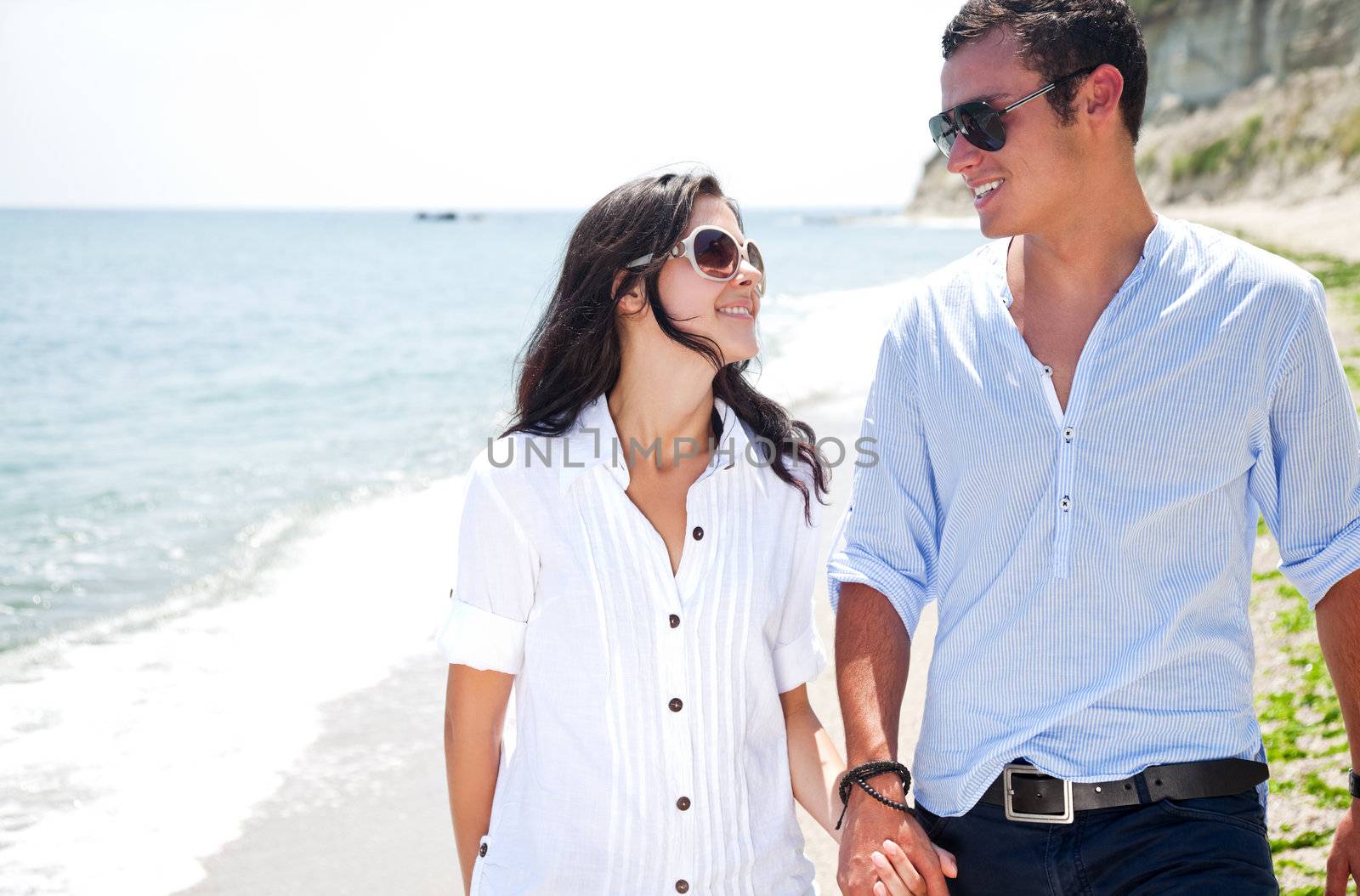 Young happy couple walking hand in hand along beach, smiling at each other