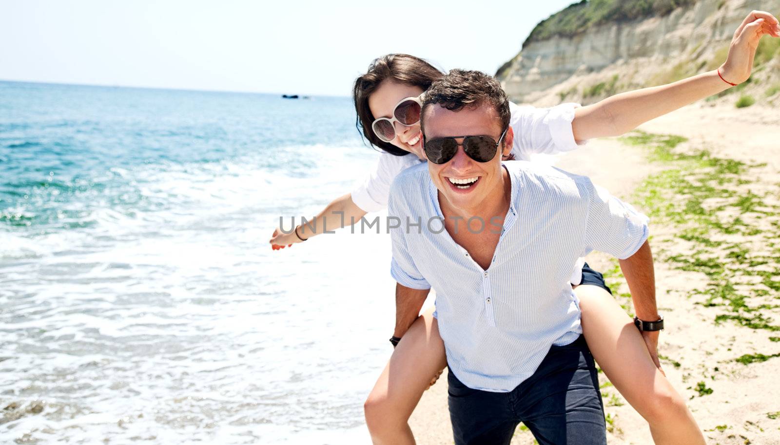 Happy young couple with sunglasses on solitary beach, man giving piggyback ride to woman