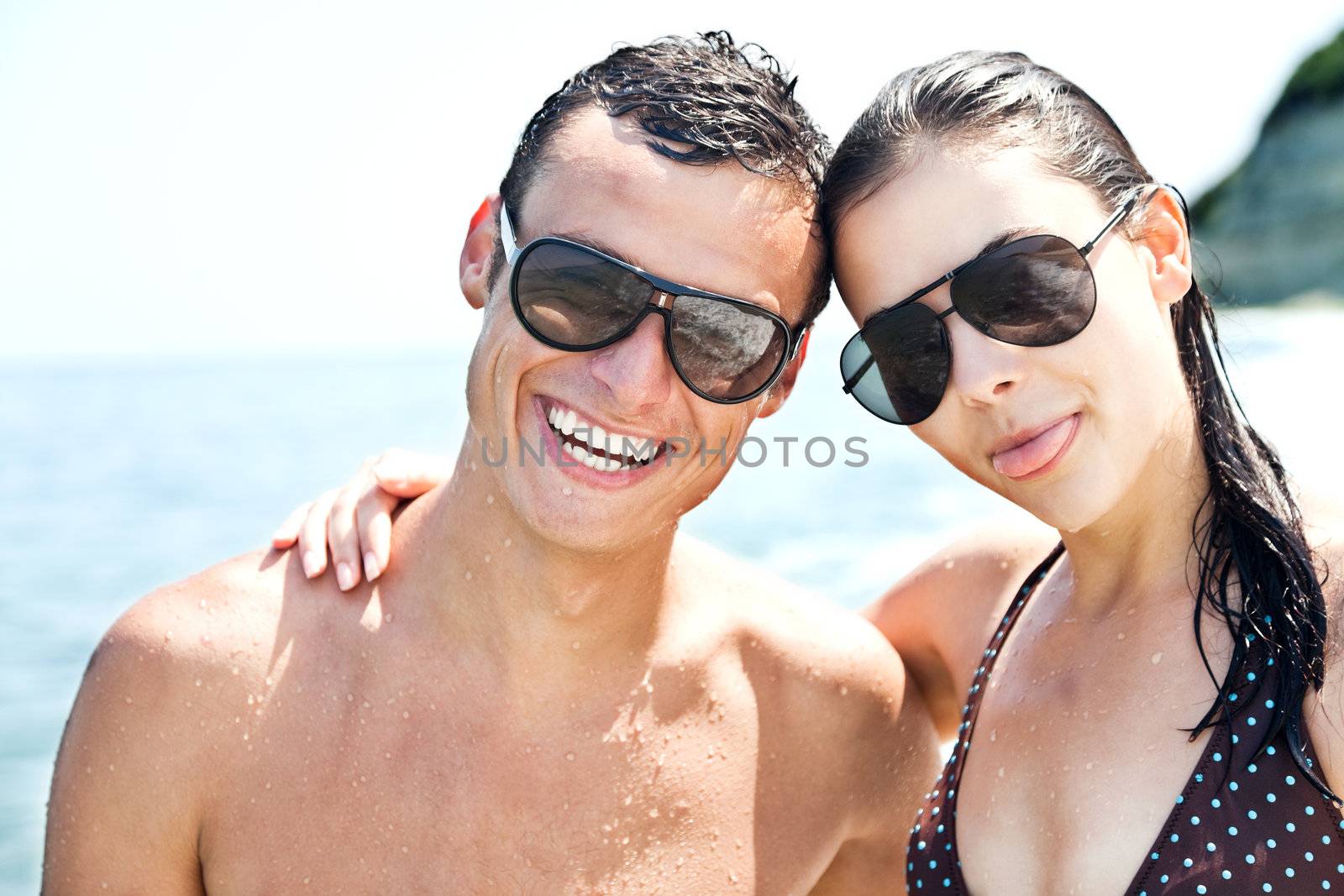 Happy couple with sunglasses hugging and smiling on beach, female with tongue out