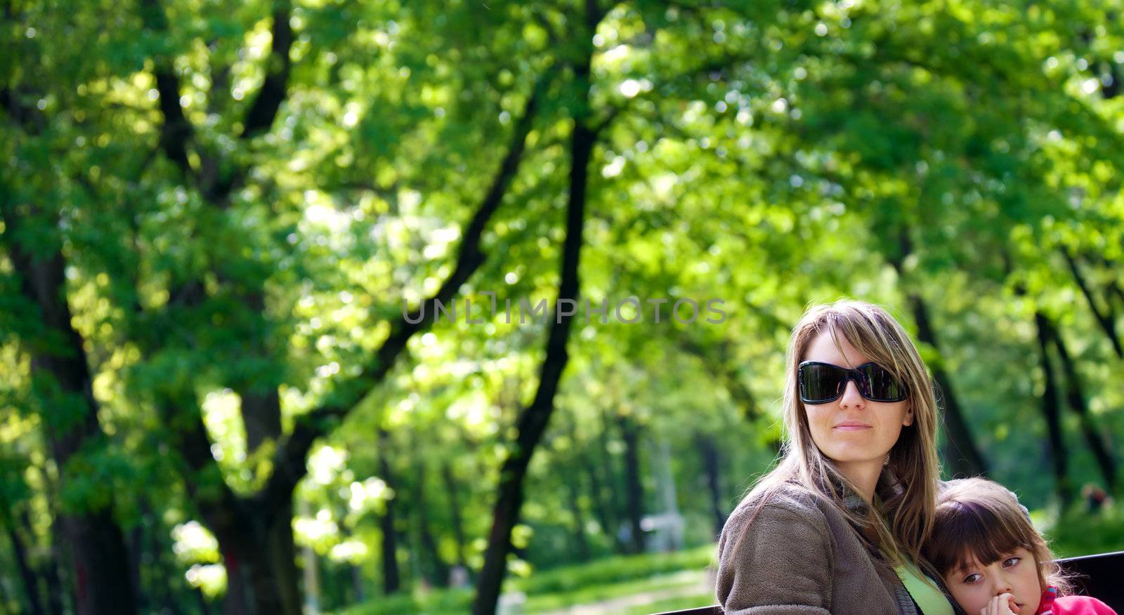 Mother with sunglasses sitting on bench in park embracing daughter