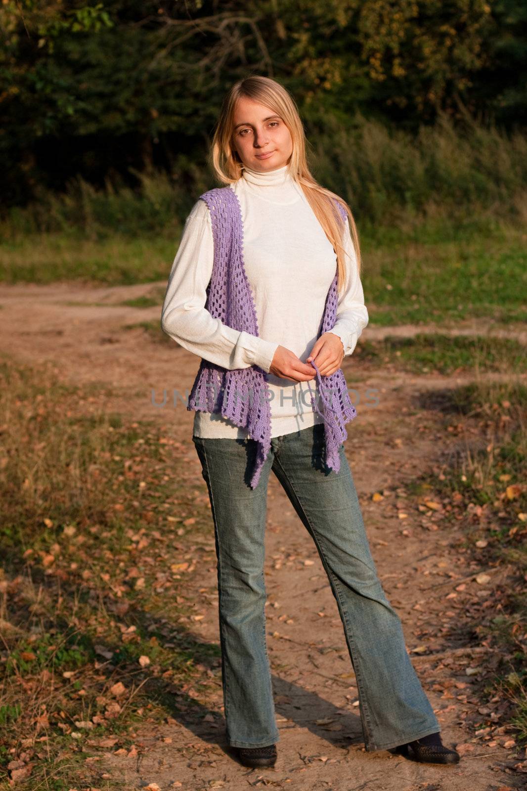 A girl in a lilac vest and white polo-neck sweater and jeans in a forest
