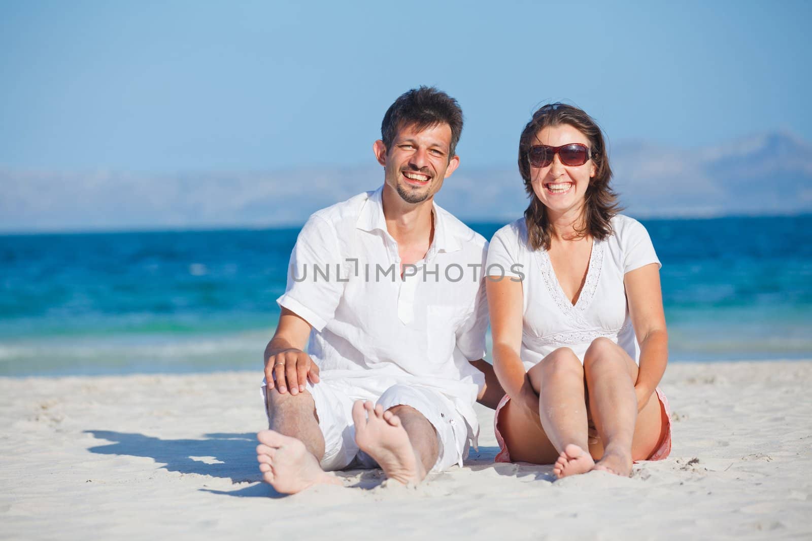 Happy young man and woman couple laughing and sitting on a deserted tropical beach with bright clear blue sky