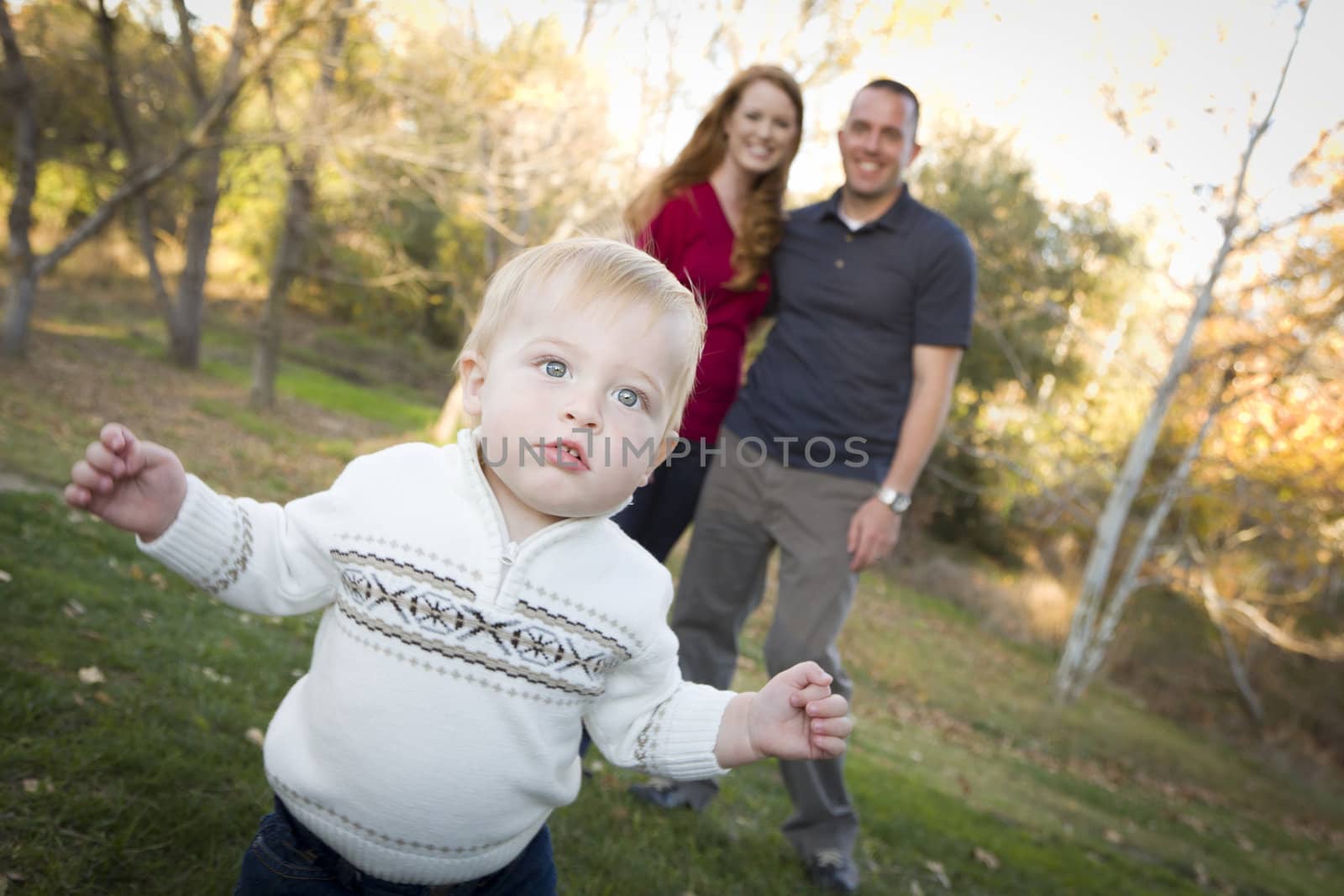 Cute Young Boy Walking in the park as Adoring Parents Look On From Behind.