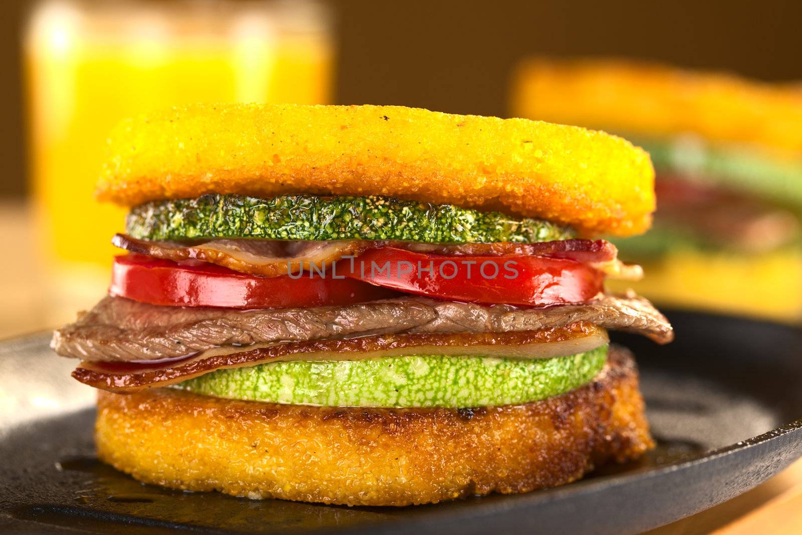 Polenta burgers with fried zucchini, bacon, beef and tomato accompanied by orange juice (Selective Focus, Focus on the front of the burger)