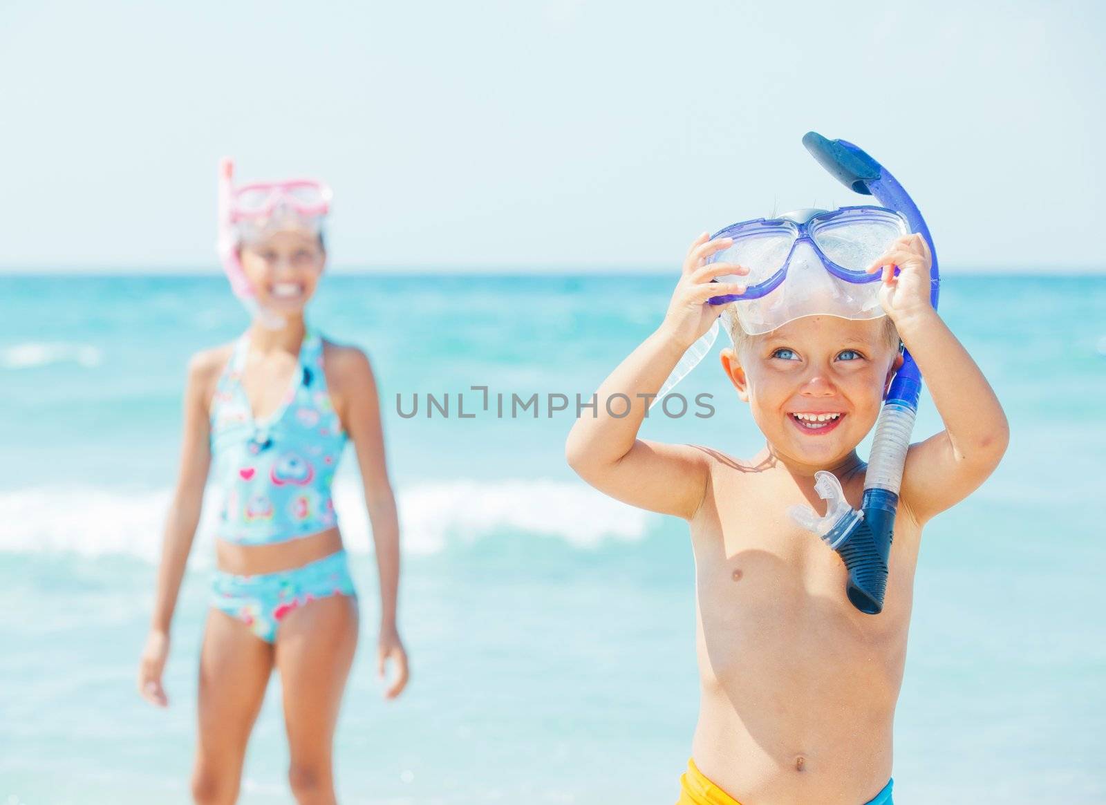 Happy young boy with snorkeling equipment on sandy tropical beach, his sister background.