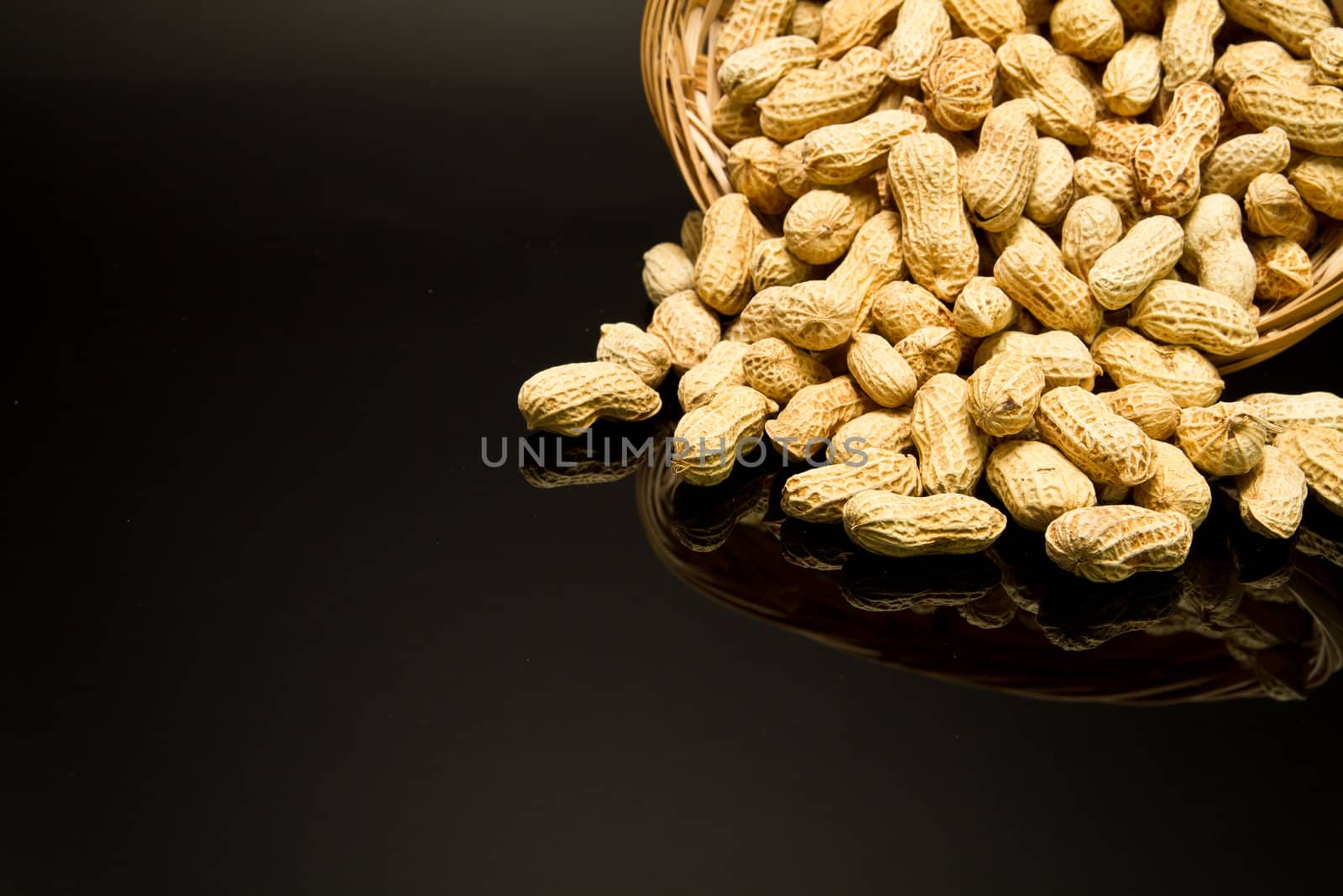 A pile of roasted groundnut spilled from a rattan basket on a black mirror with mirror image off-centered to the top right leaving empty space for text