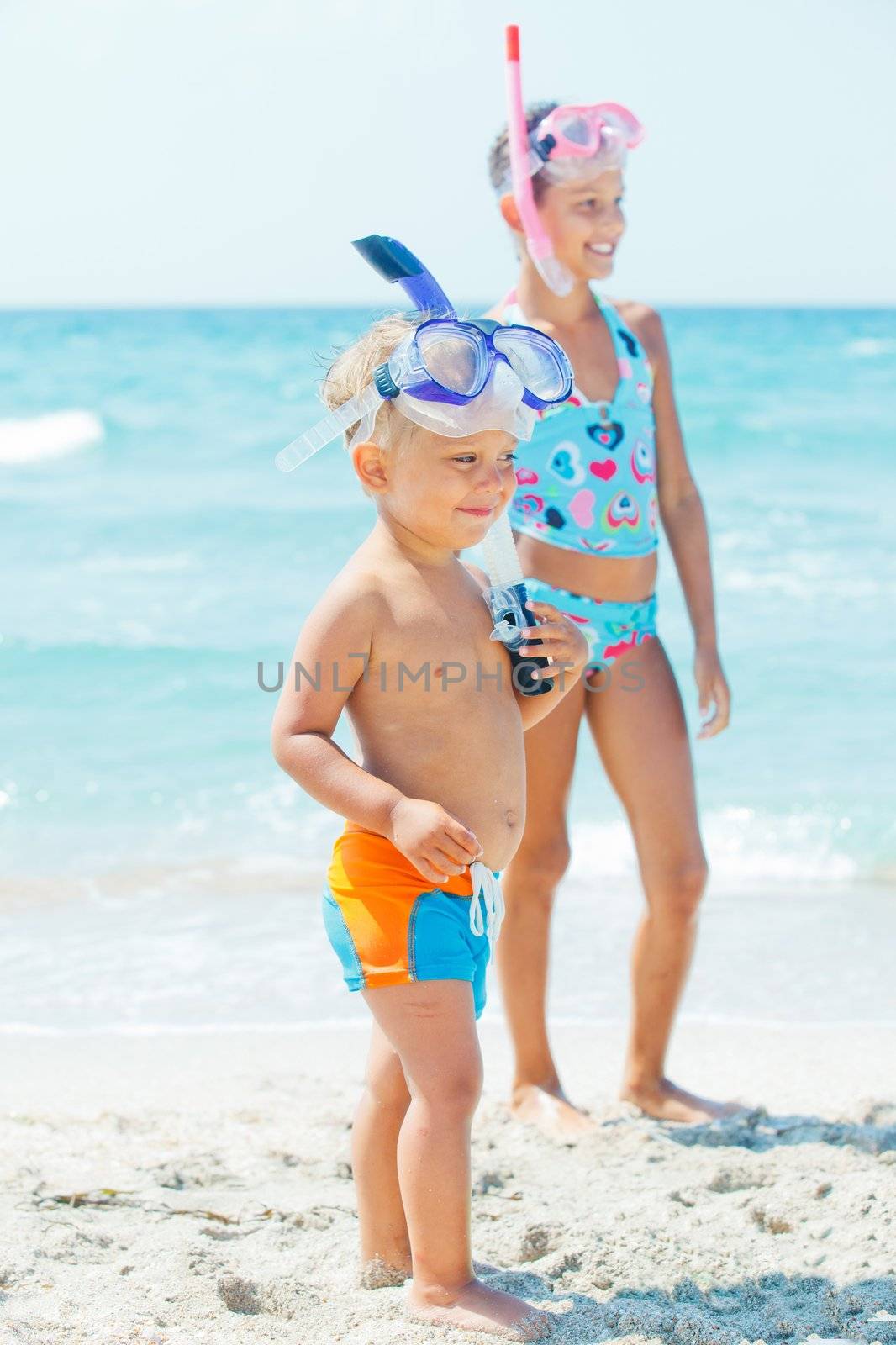 Happy young boy with snorkeling equipment on sandy tropical beach, his sister background.