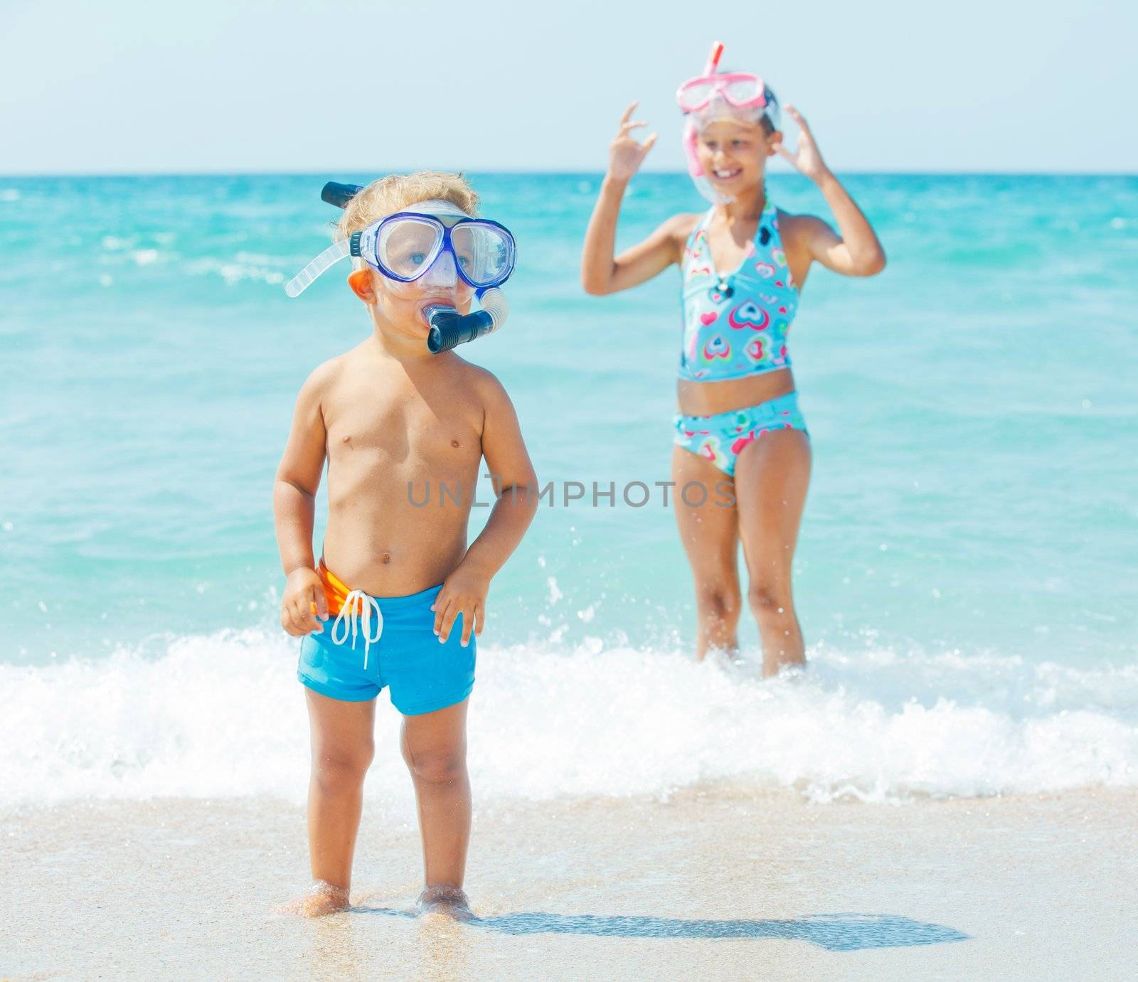 Happy young boy with snorkeling equipment on sandy tropical beach, his sister background.