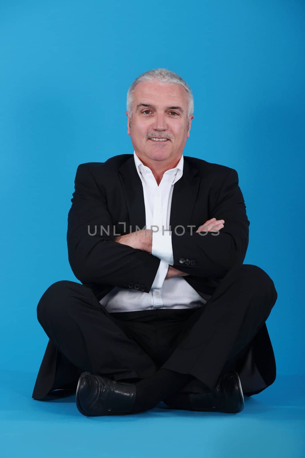 mature man sitting cross-legged and cross-armed against blue background