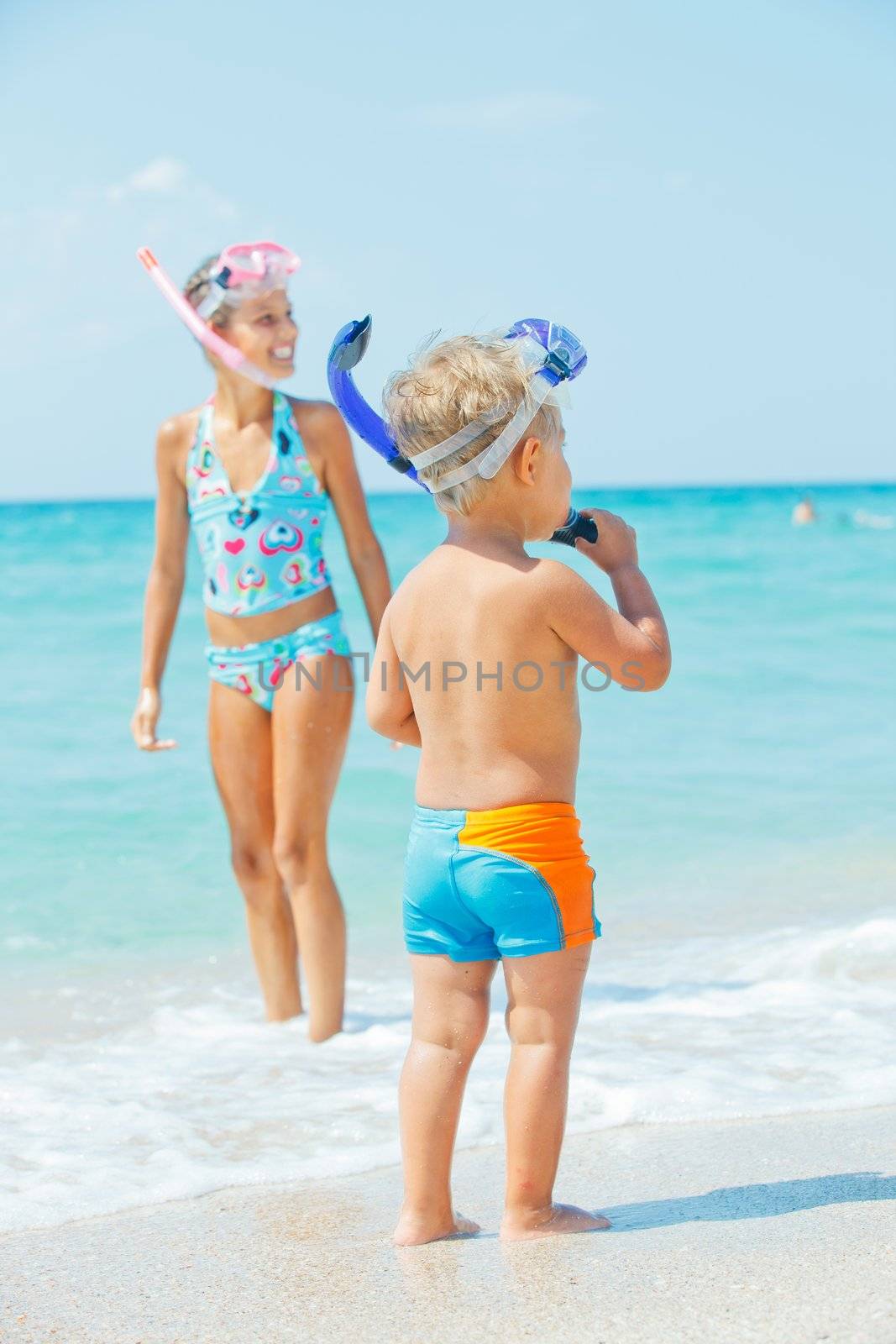 Happy young boy with snorkeling equipment on sandy tropical beach, his sister background. Vertical view