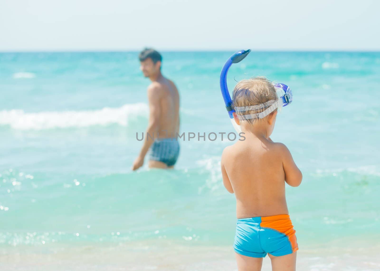 Cute little boy with snorkeling equipment on tropical beach, his father background.