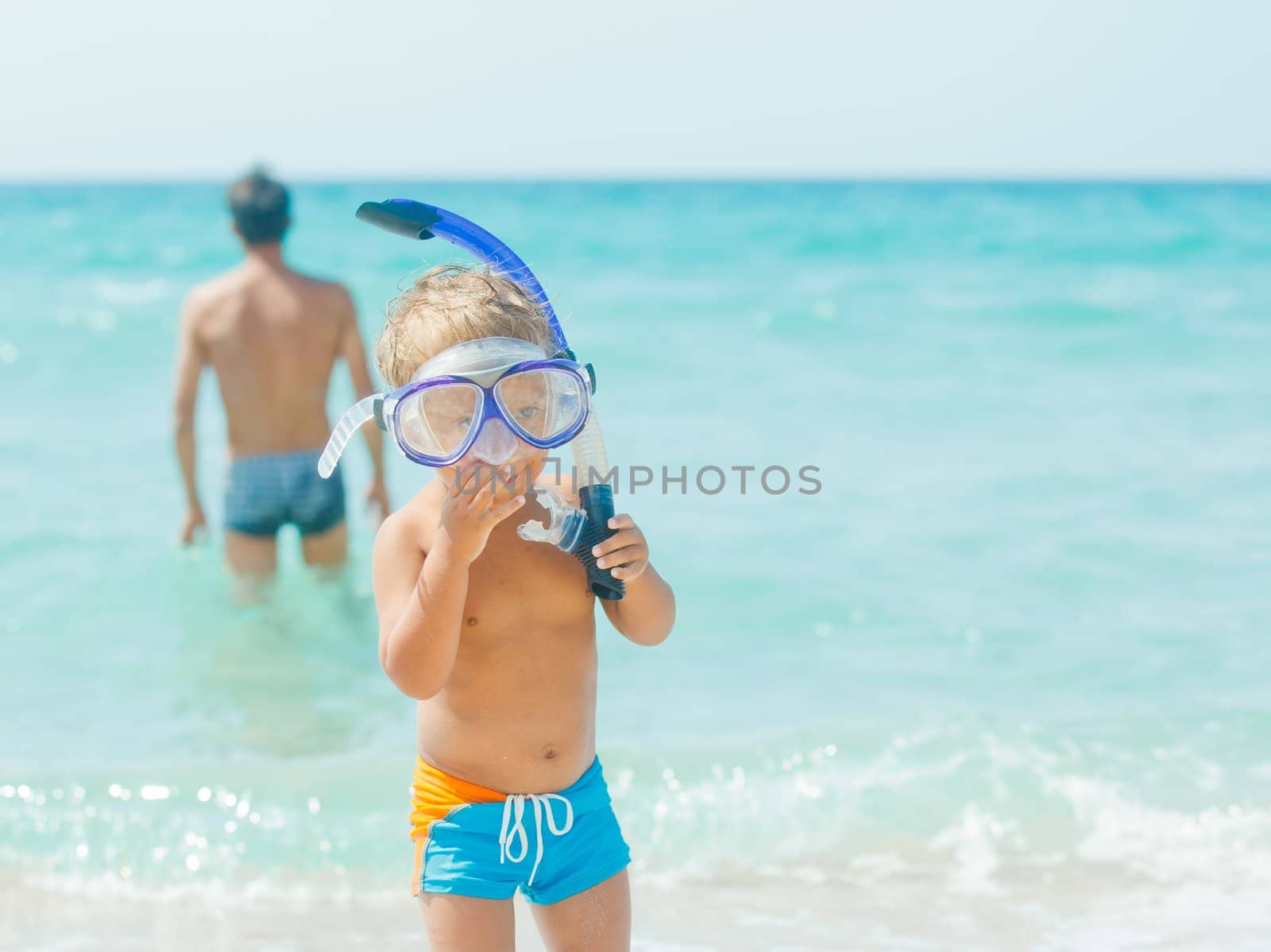 Cute little boy with snorkeling equipment on tropical beach, his father background.