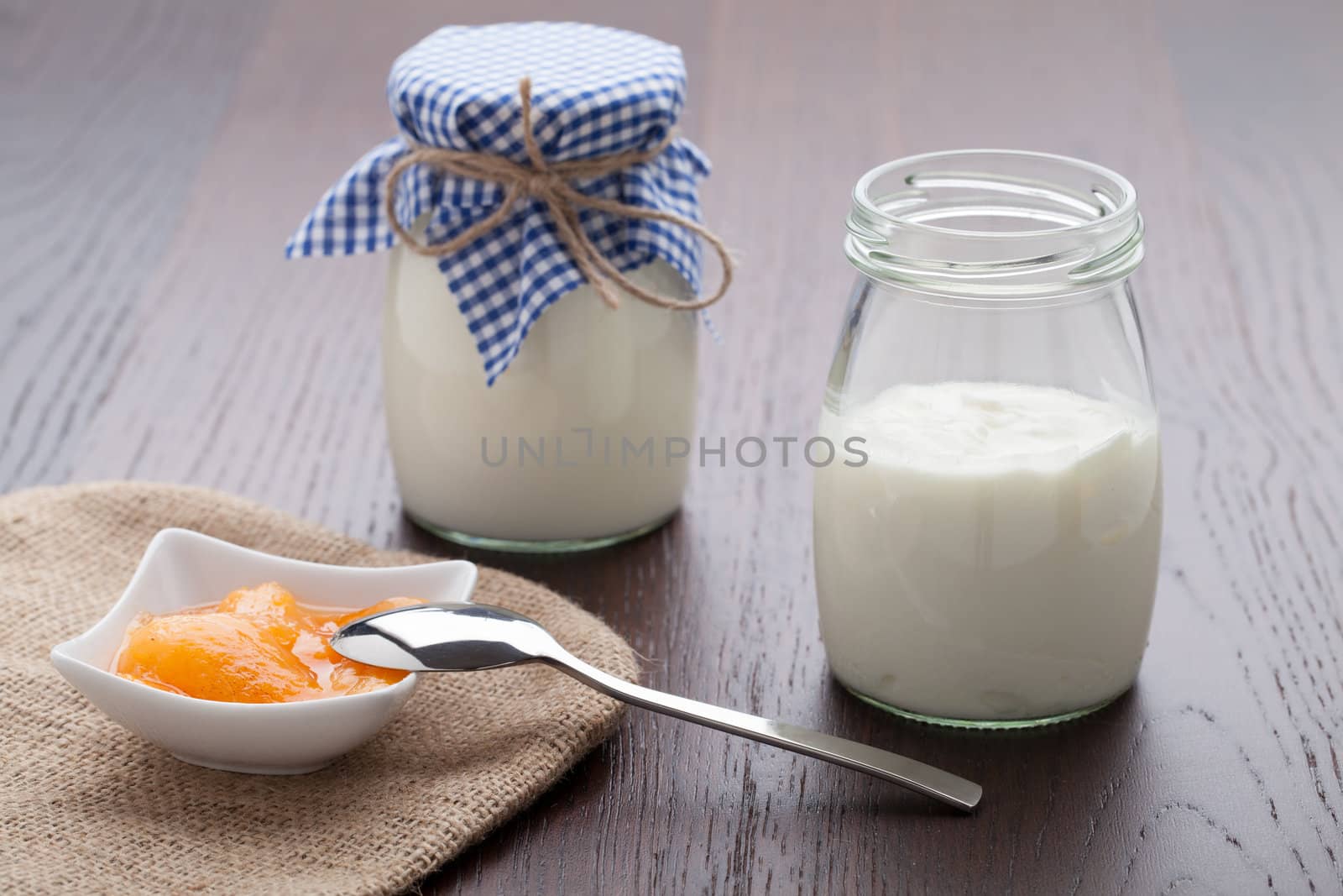 Homemade milk yogurt with peach or apricot jam in porcelain bowl and metal spoon served on linen napkin, wooden table