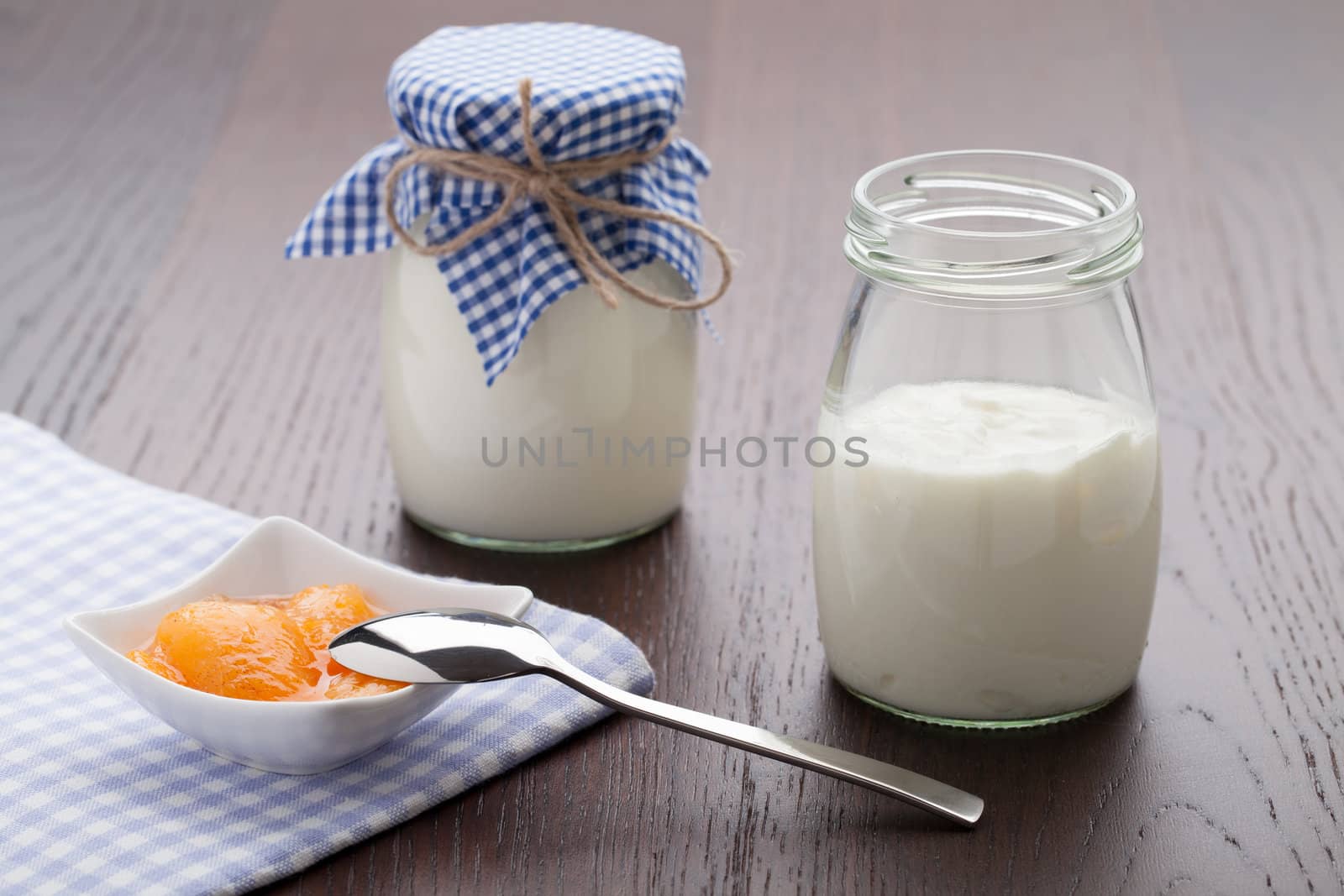 Homemade milk yogurt with peach or apricot jam in porcelain bowl and metal spoon served on linen napkin, wooden table