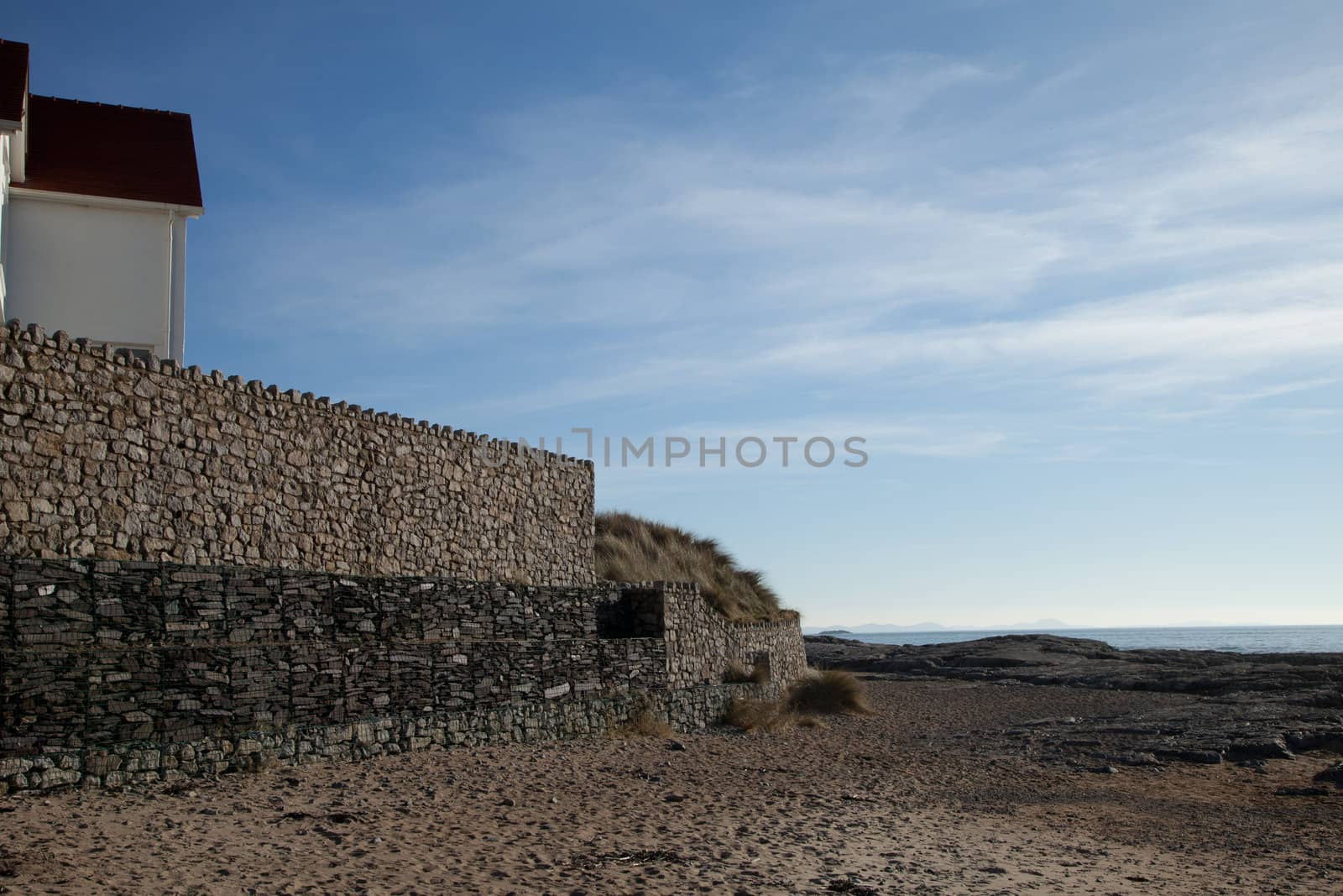 A high solid stone wall with cubed crates of stone reinforcements at sand level on a beach.