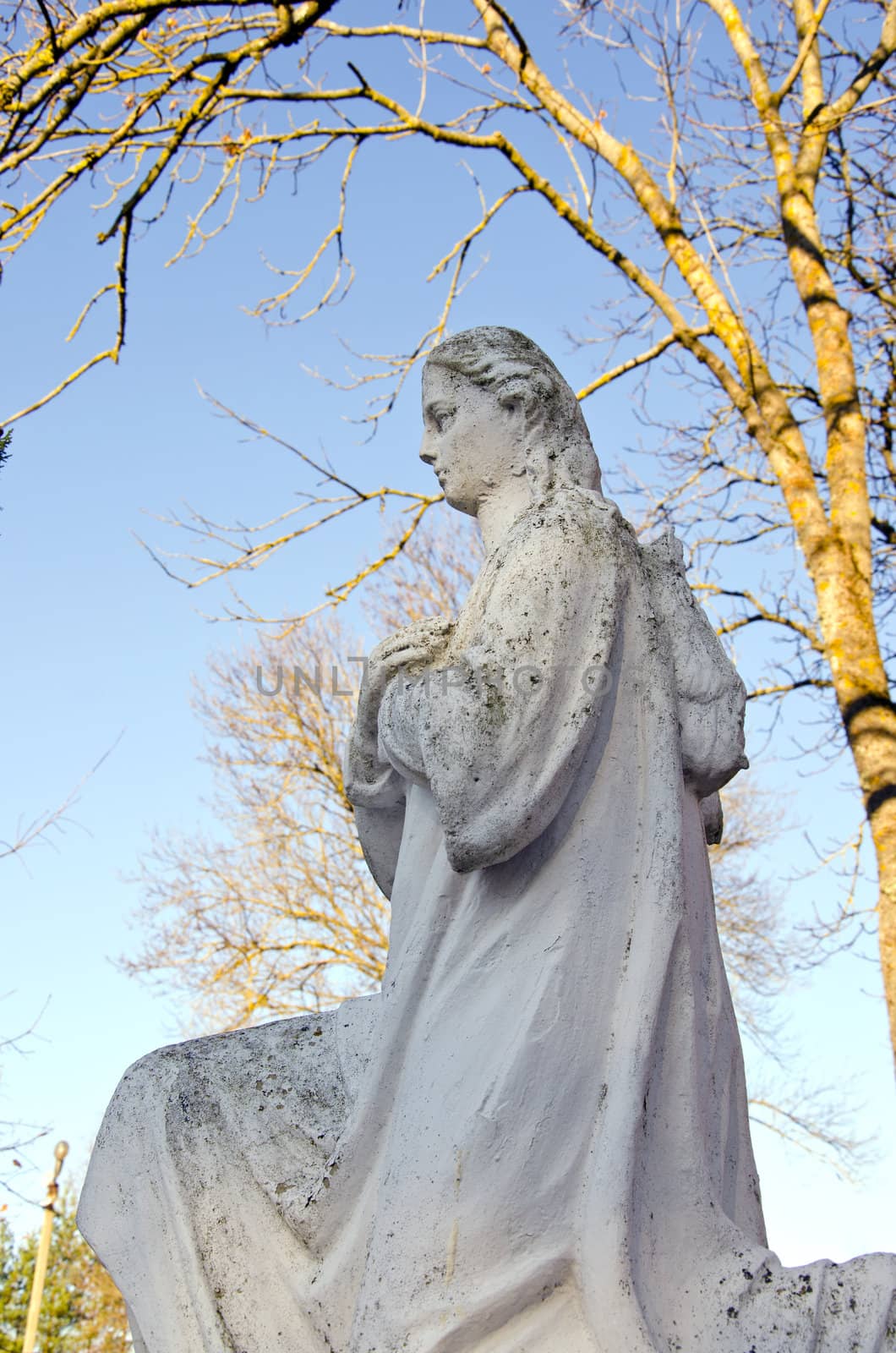 Dirty old white angel statue without wings. Trees without leaves in autumn.