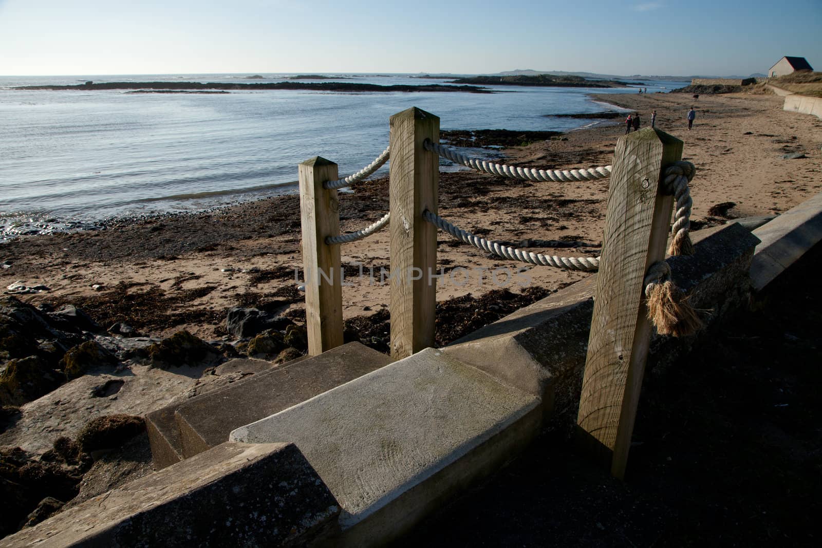 Concrete set steps with wooden posts and a rope hand rail leads to a beach with people in the distance.