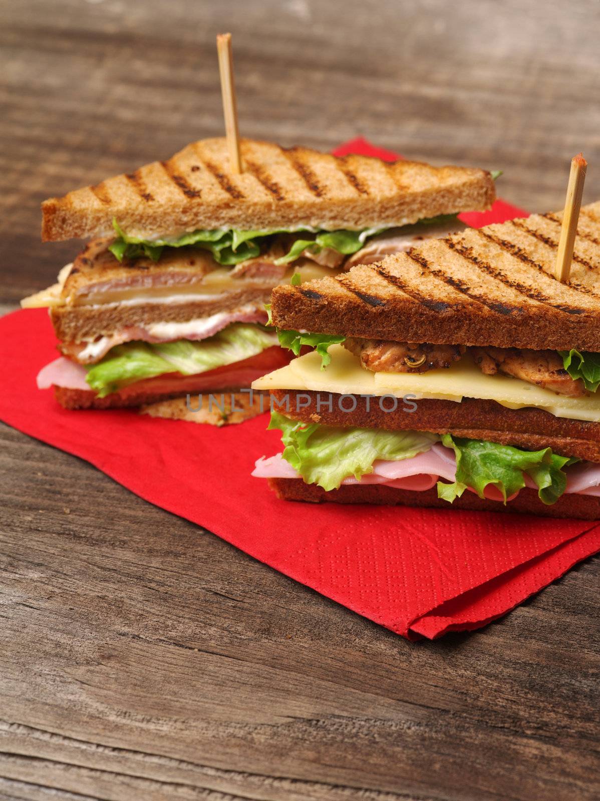 Photo of a club sandwich made with turkey, bacon, ham, tomato, cheese, lettuce on a red napkin and old wood picnic table.
