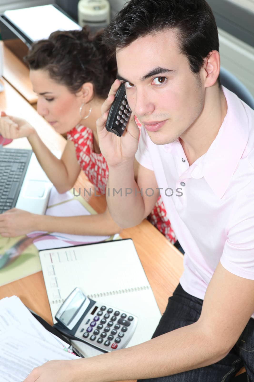 Young man on the phone whilst sitting on an office desk by phovoir