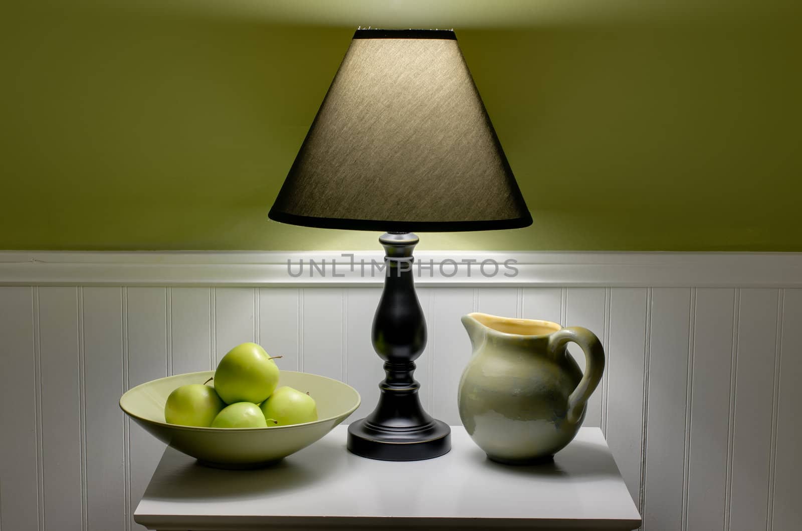 Bowl of green apples, lamp and pitcher on table.  Scene is illuminated only by soft light from lamp.