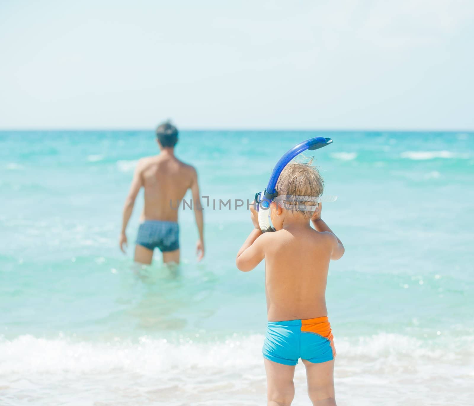 Cute little boy with snorkeling equipment on tropical beach, his father background.