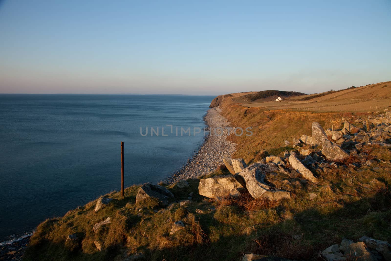 Stones and a post in the red evening light with steep mud and grass banks leading to a pebble shore and the sea.