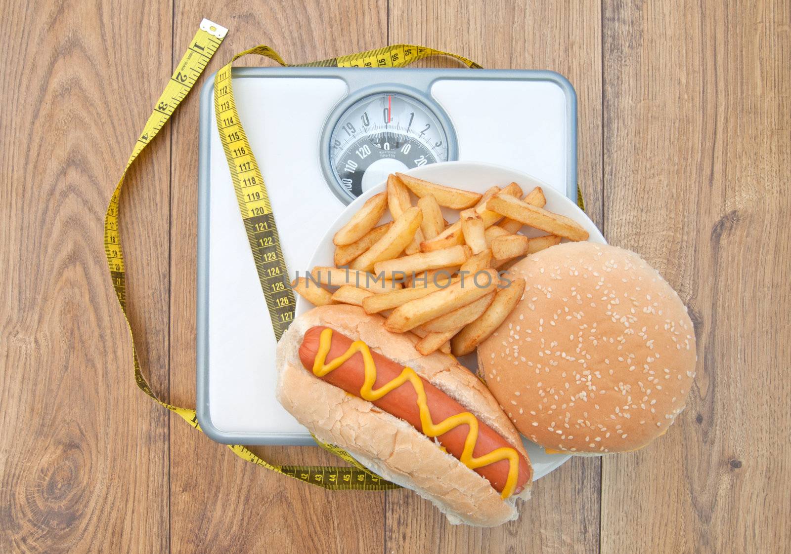 Fattening foods on weighing scales with a tape measure, including burger chips and hot dog