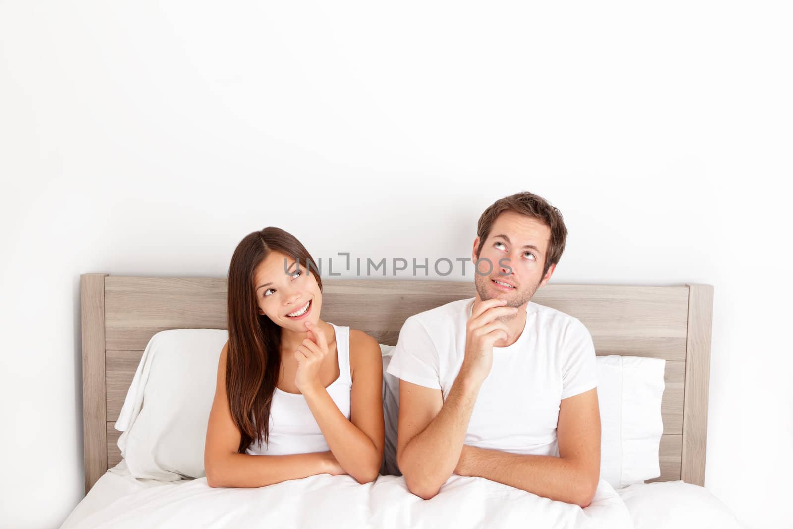 Thinking young couple sitting pensive in bed smiling and looking up at the ceiling for inspiration and ideas. Happy young interracial couple, Asian woman, Caucasian man.