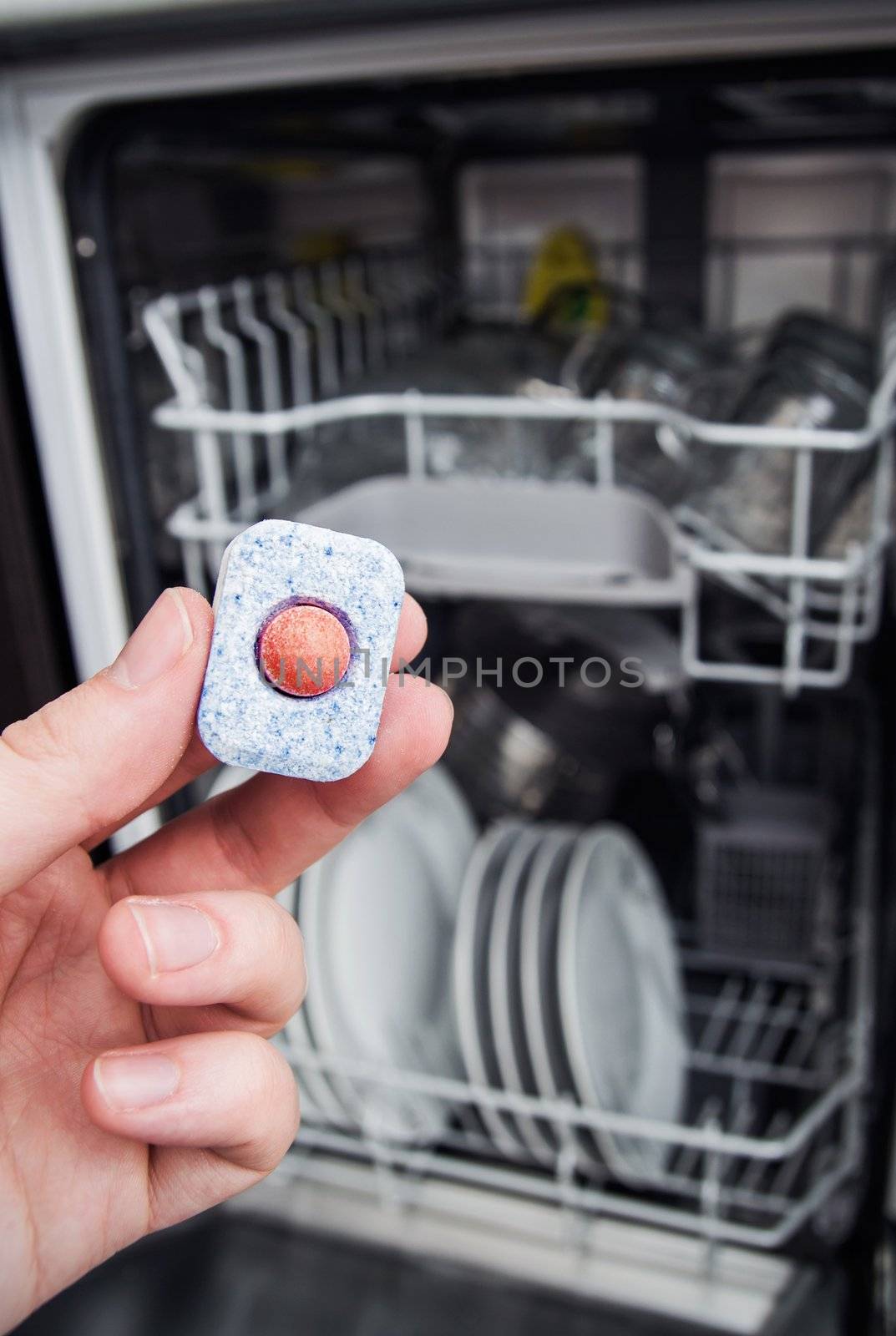 Hand holding dishwashing tablet. Dishwasher in background