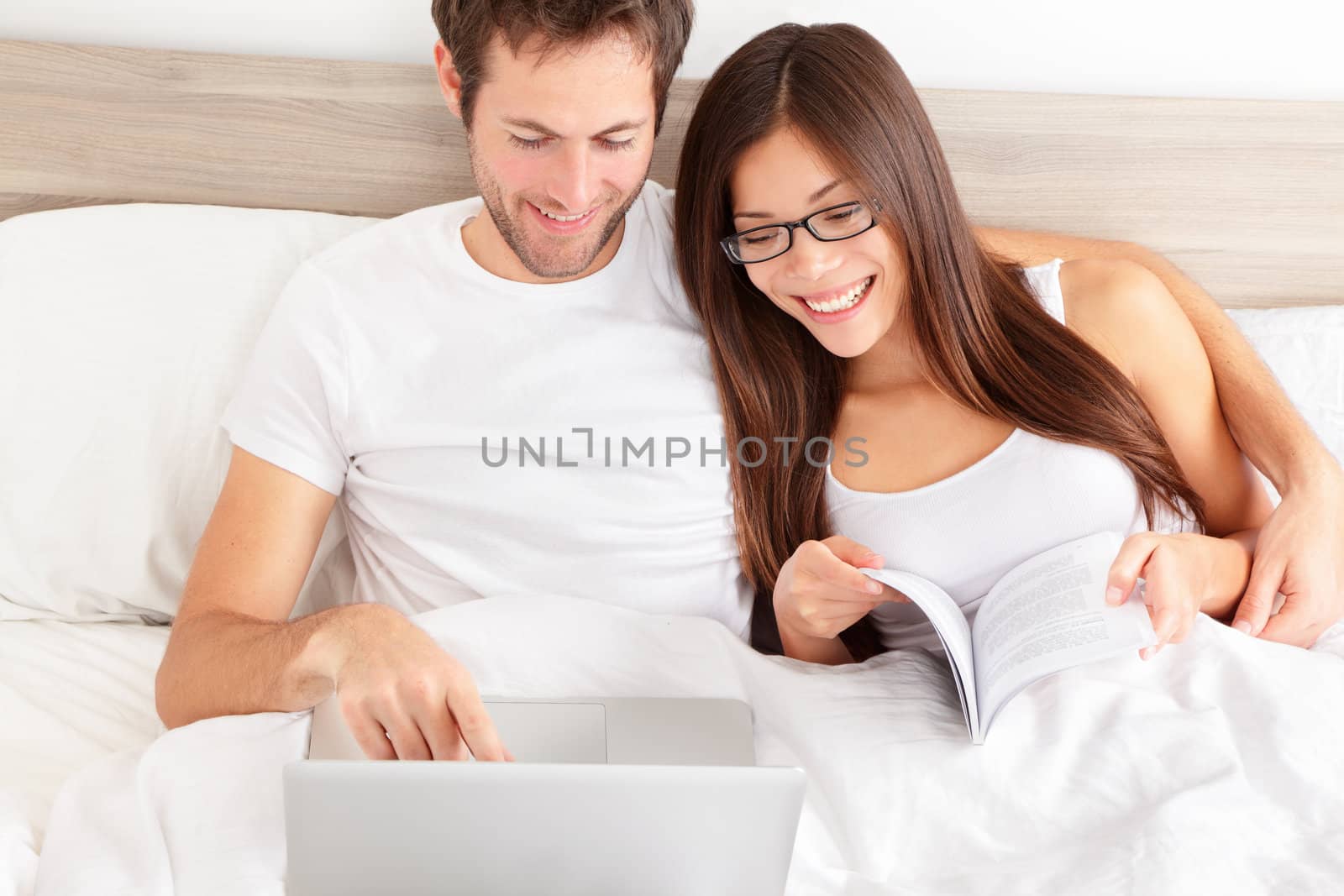 Happy affectionate young married couple sitting side by side in bed relaxing and laughing at information on the screen of their laptop. Happy young interracial couple, Asian woman, Caucasian man.