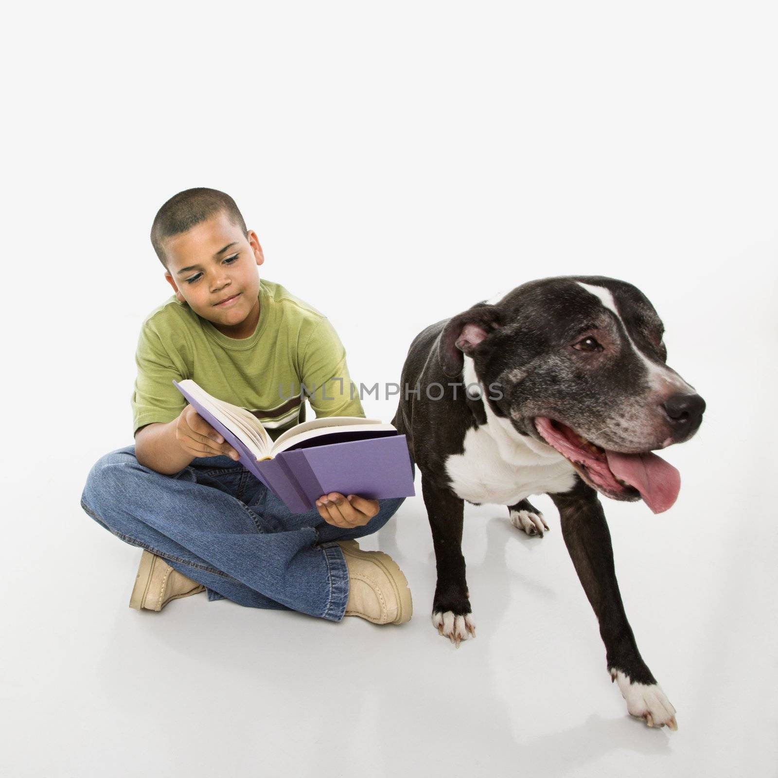 Young hispanic boy reading book while his dog sits nearby.