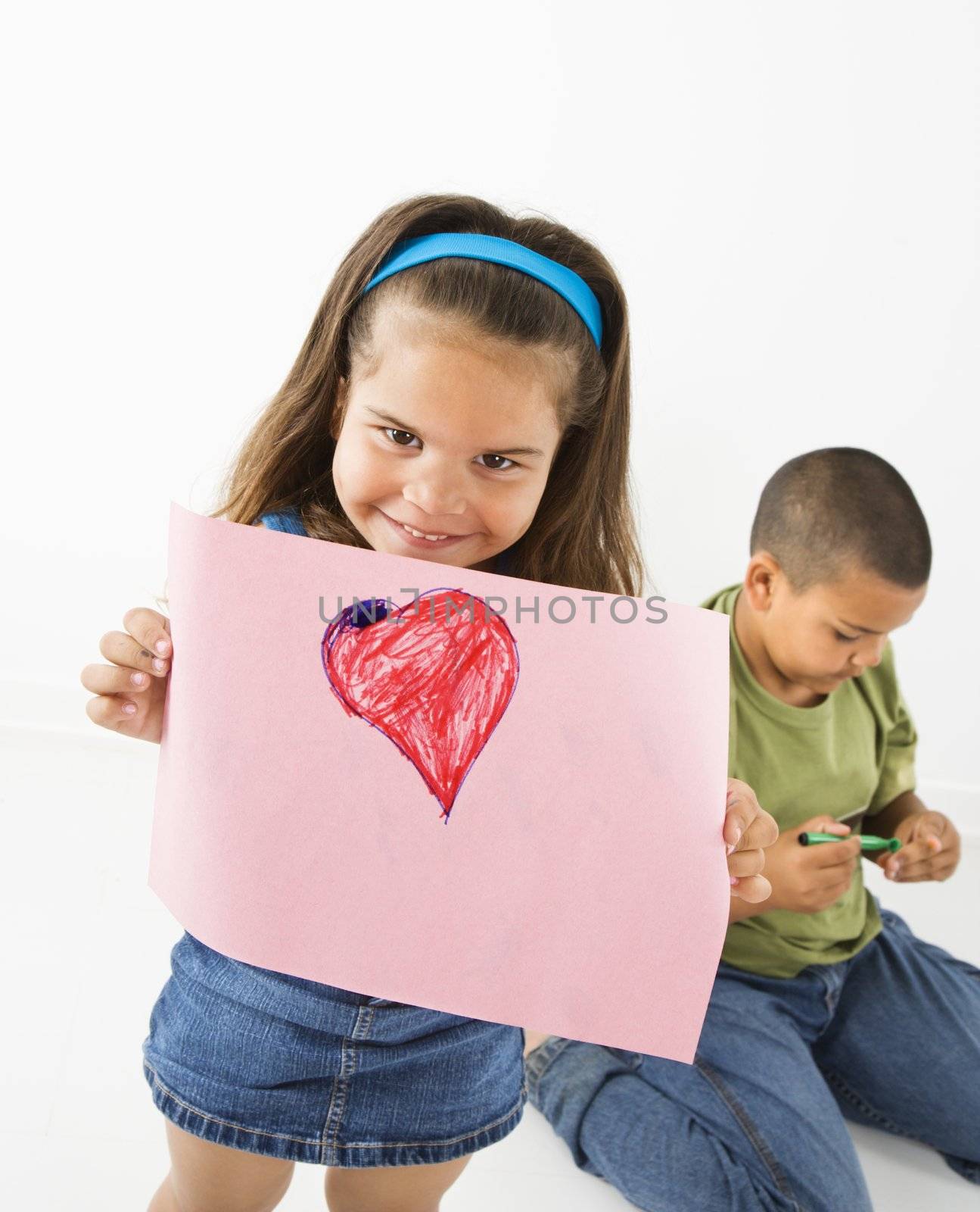 Young girl showing off drawing of heart while boy sits behind her.
