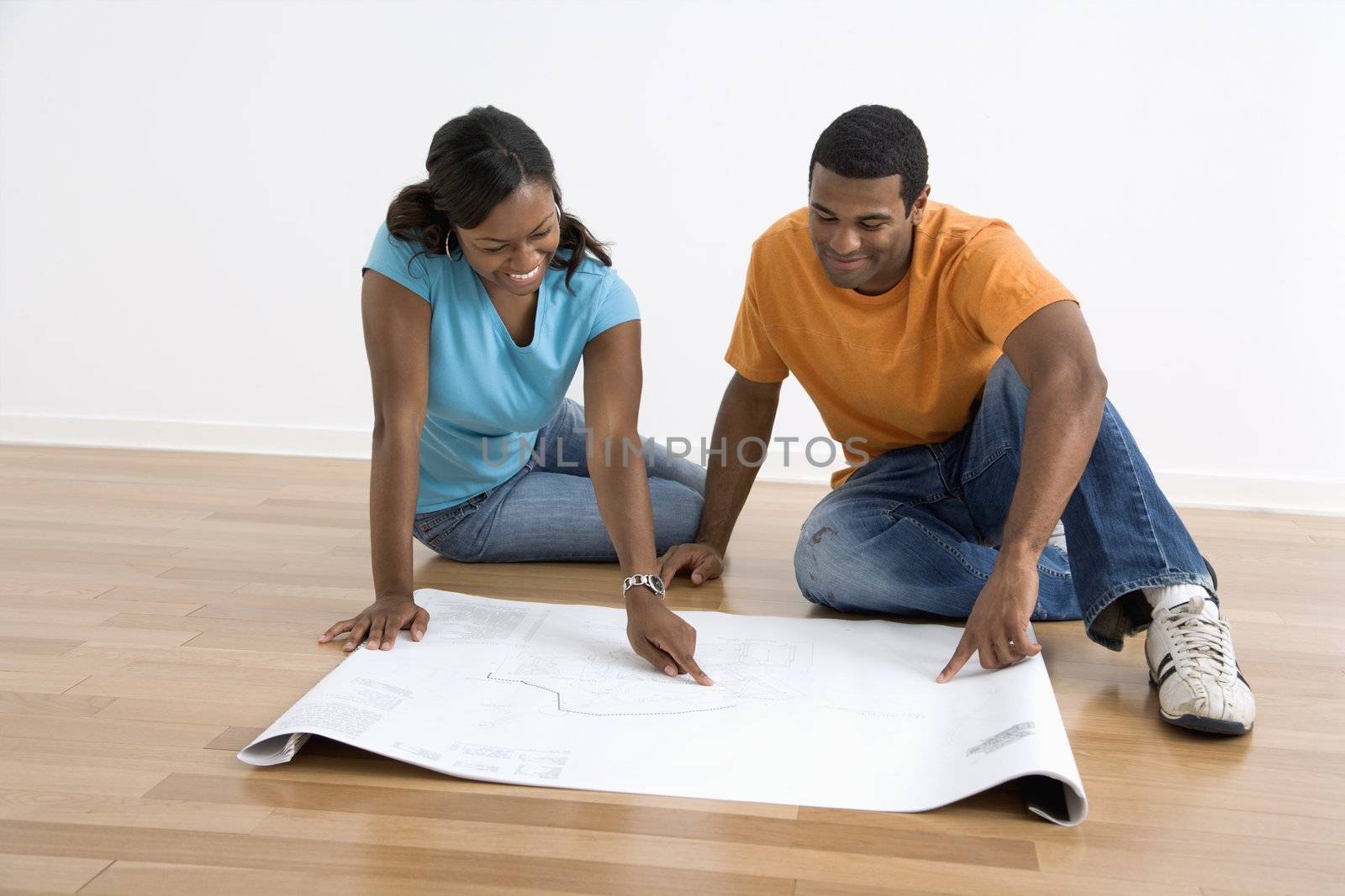 African American male and female couple sitting on floor looking at architectural blueprints.