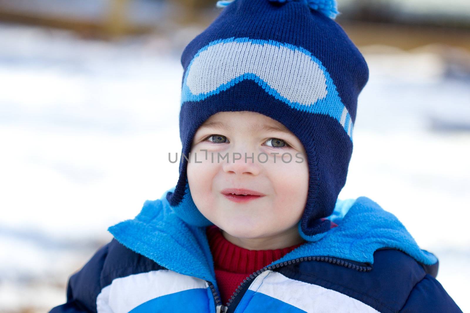 The photograph is a portrait of a very cute, handsome little boy playing in the snow.  He is wearing a blue coat and hat and has a mischievous expression on his face.