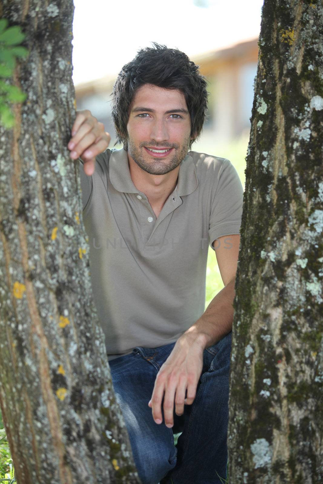 25 years old man crouching on his heels behind two trees