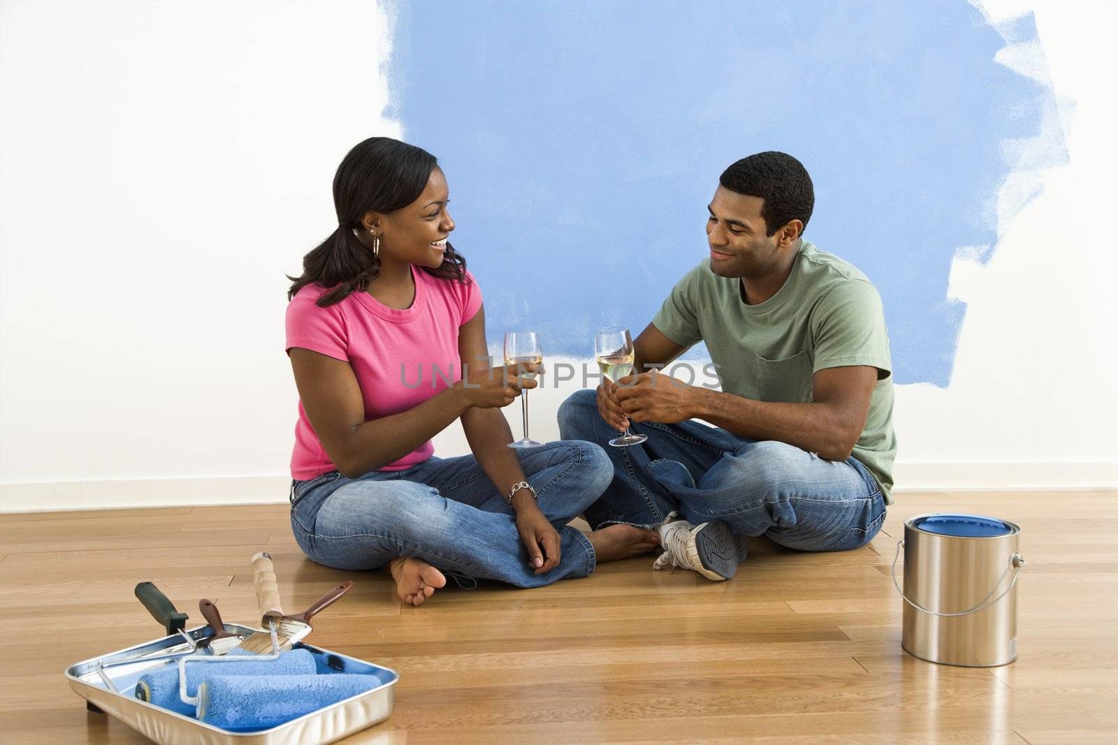 African American couple relaxing together with wine next to half-painted wall and painting supplies.