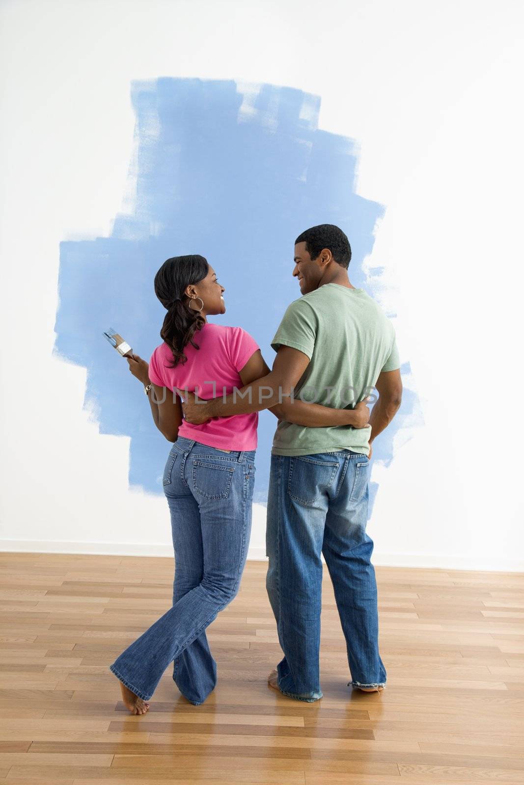 African American couple standing together next to half-painted wall discussing their work.