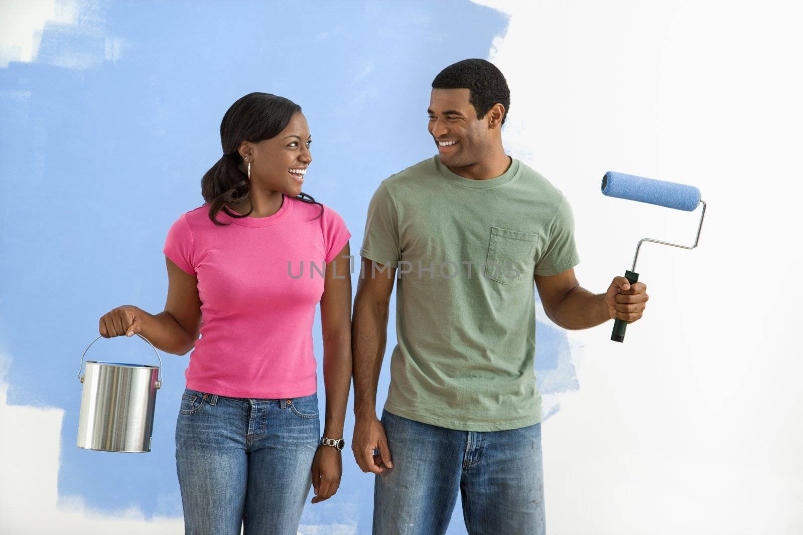 African American smiling couple next to half-painted wall with paint supplies.