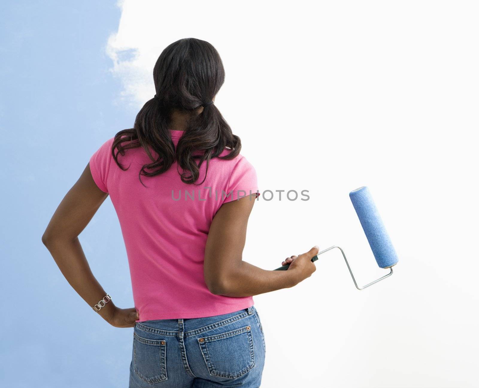 African American young woman standing next to half-painted wall with paint roller.