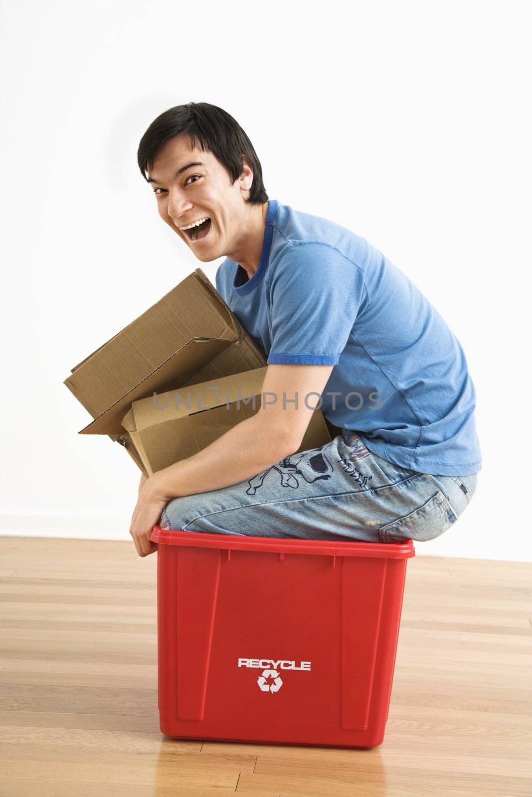 Asian young man trying to sit in recycling bin with cardboard.